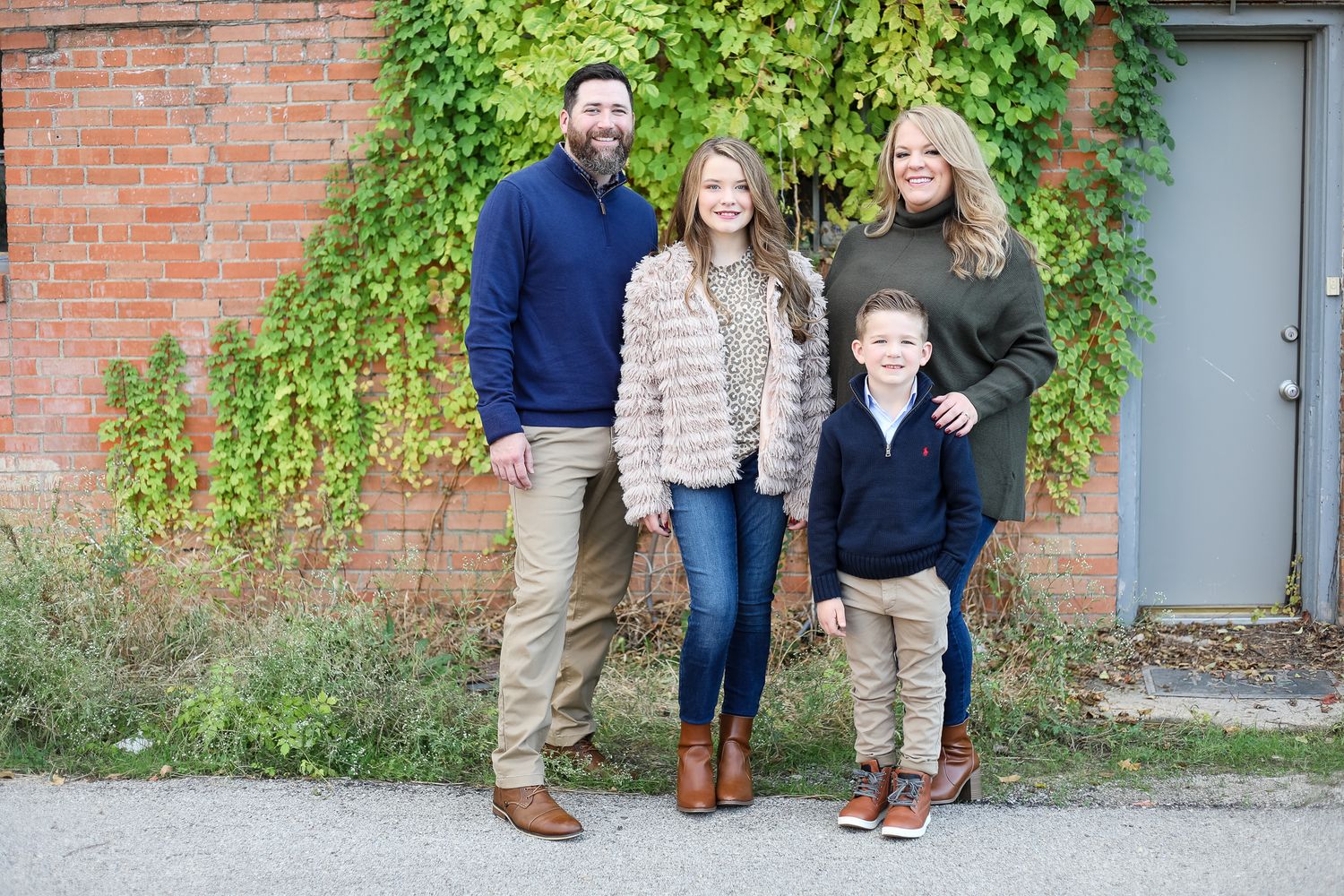 A family of four stands together outdoors against an ivy-covered red brick wall on an autumn day.