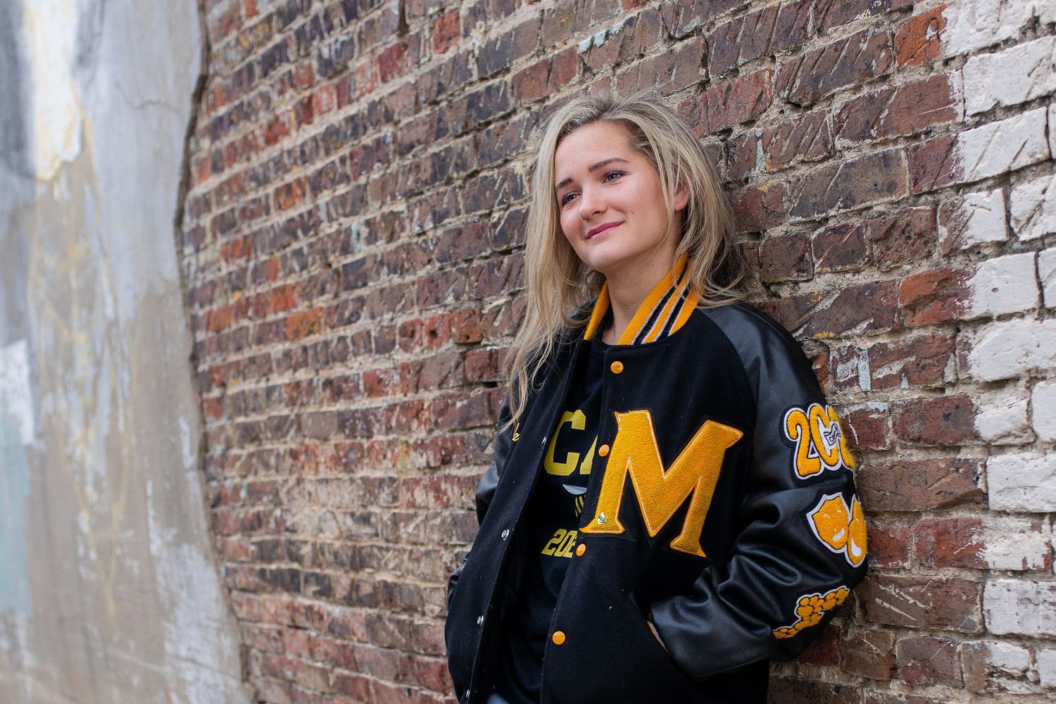 A person wearing a black and gold varsity letterman jacket poses against a rustic brick wall.