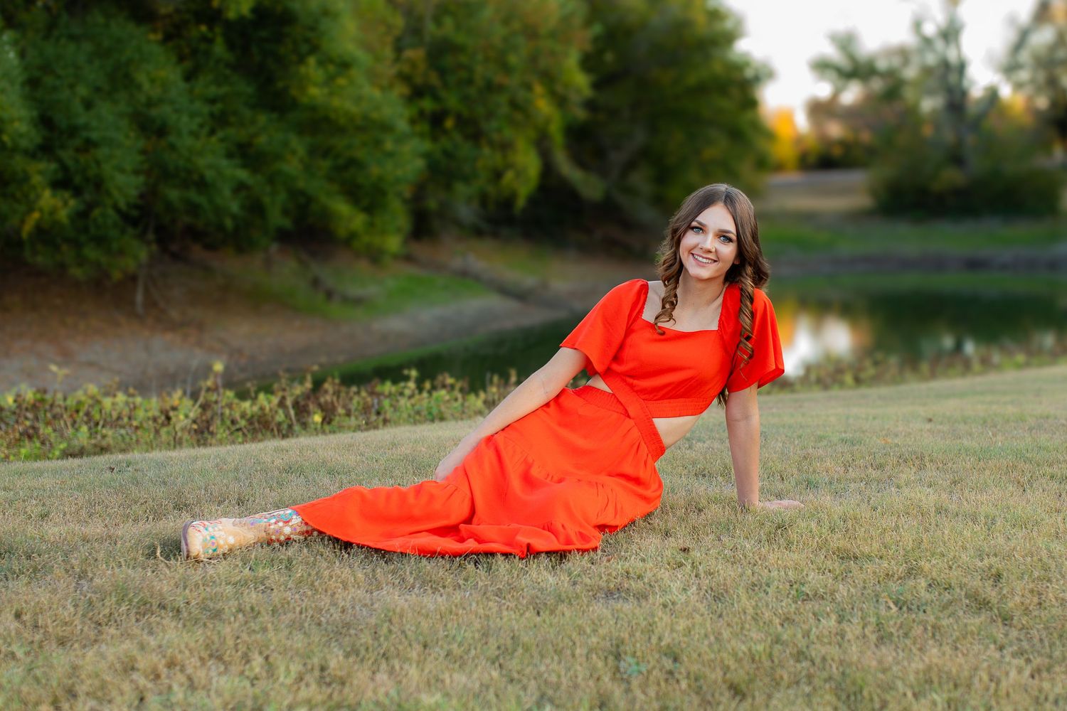 A woman in a flowing red dress reclines on grassy ground near a tranquil pond at sunset.
