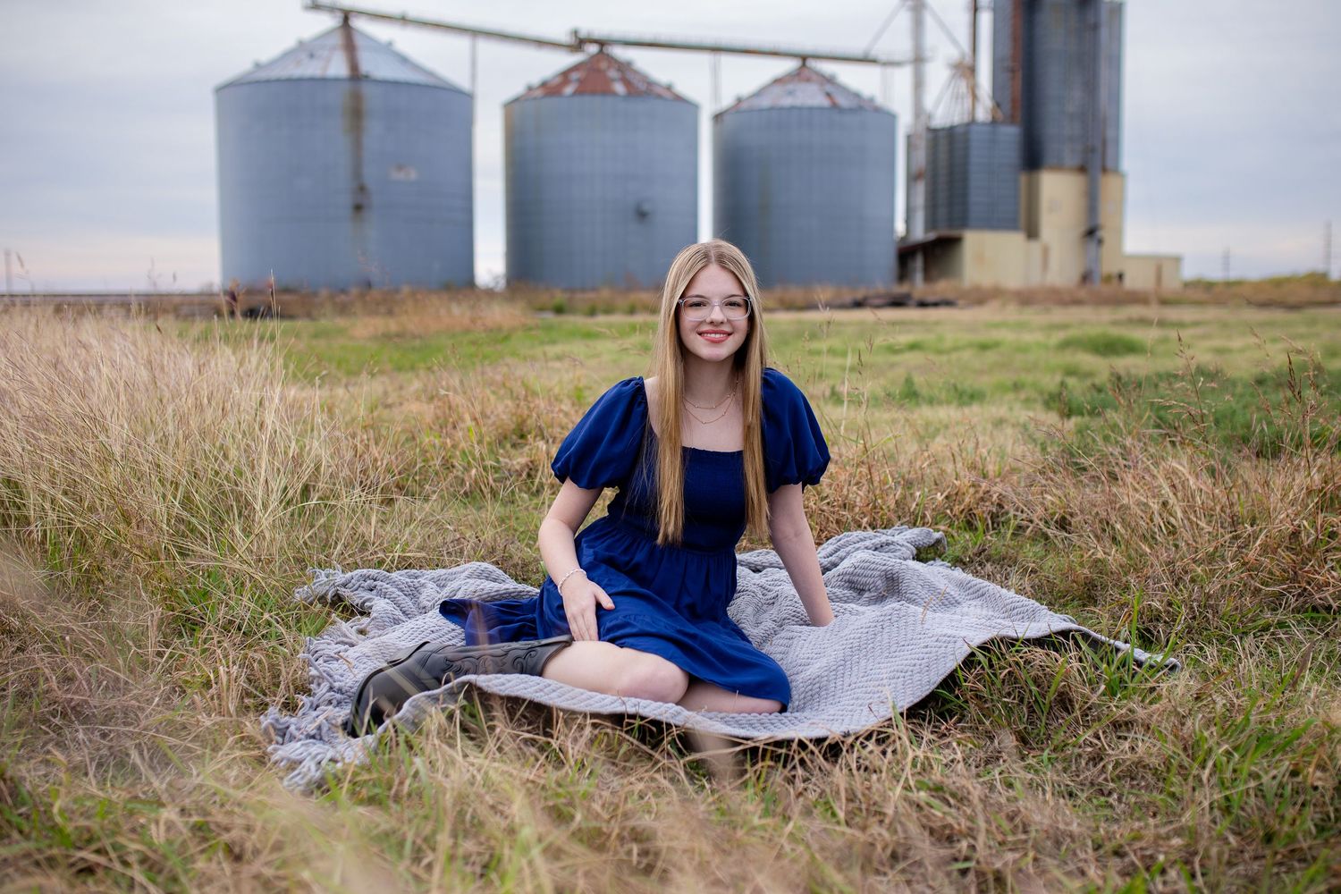 A woman in a blue dress lies gracefully on a blanket in a rural field with grain silos and tall grass in the background.