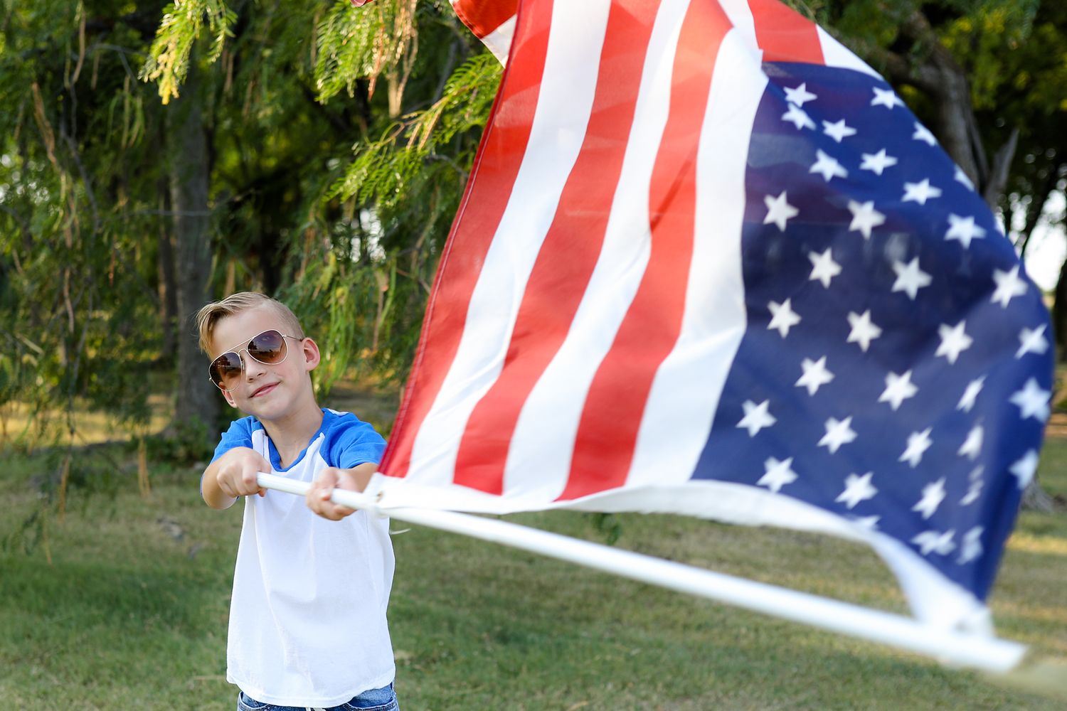 Figure waving American flag outdoors on sunny day with trees in background.