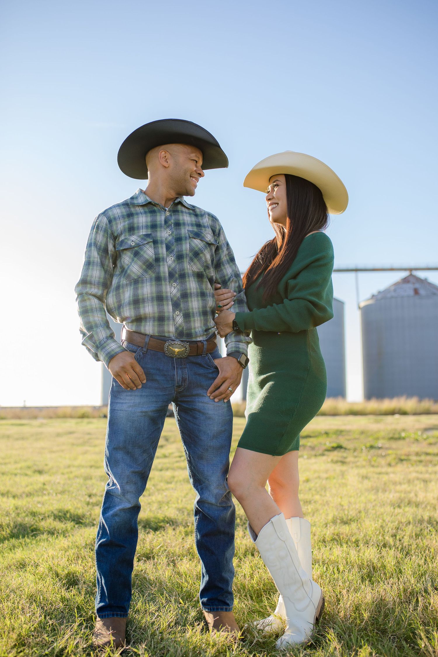Two people in western attire stand together in front of grain silos on a grassy field during sunset.