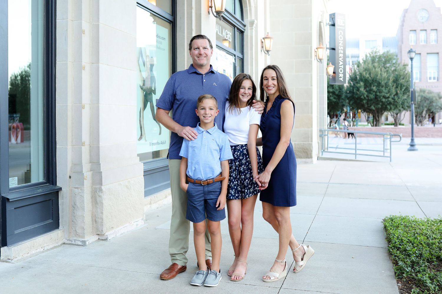Family of four poses together on downtown city sidewalk outside storefront on sunny day.