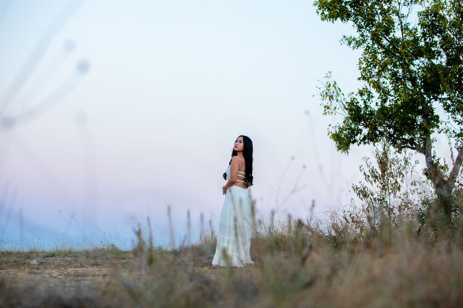 A person in a white dress stands in a field at dusk with trees in the background.