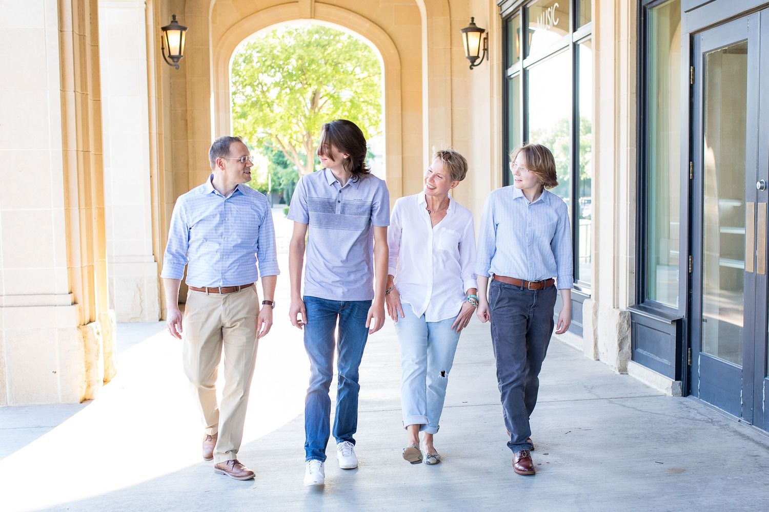 A group of people walking together in casual attire through a covered outdoor corridor with arched windows.