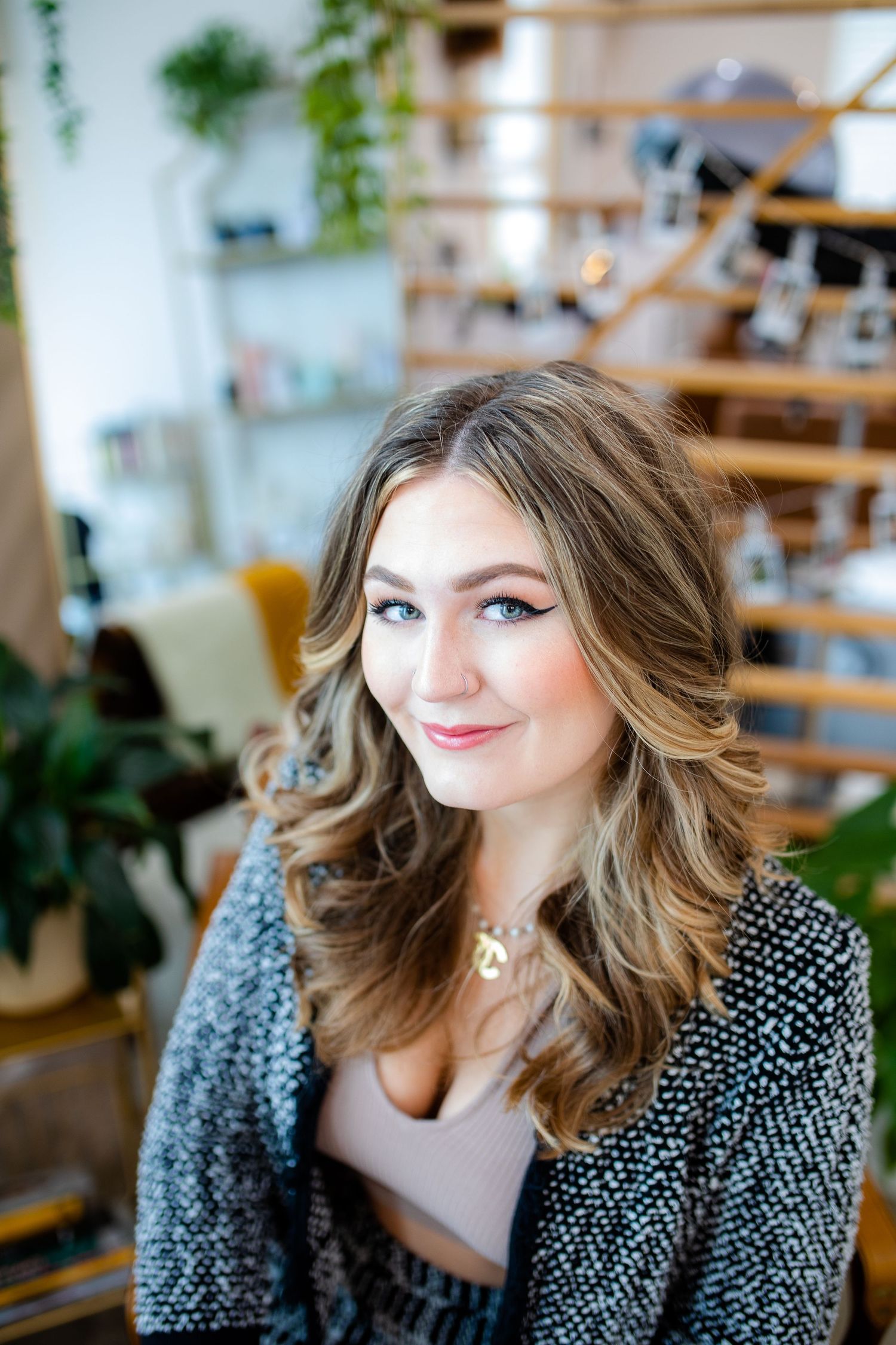 Portrait in a cozy room with wooden shelves and plants, wearing a textured grey cardigan over a neutral top.