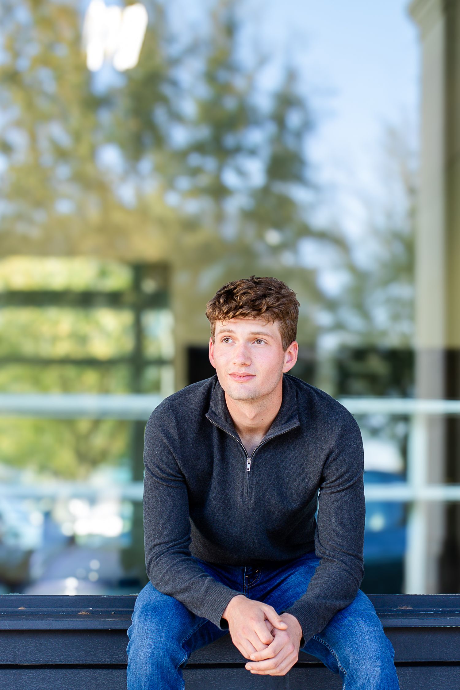 Young person in a dark quarter-zip sweater and jeans sits thoughtfully on a bench outdoors with trees and buildings in background.