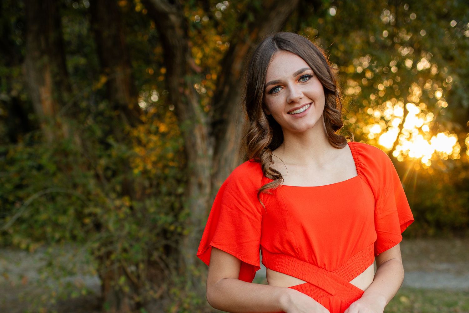 Portrait of someone in bright red dress standing outdoors during golden hour with autumn trees in background.