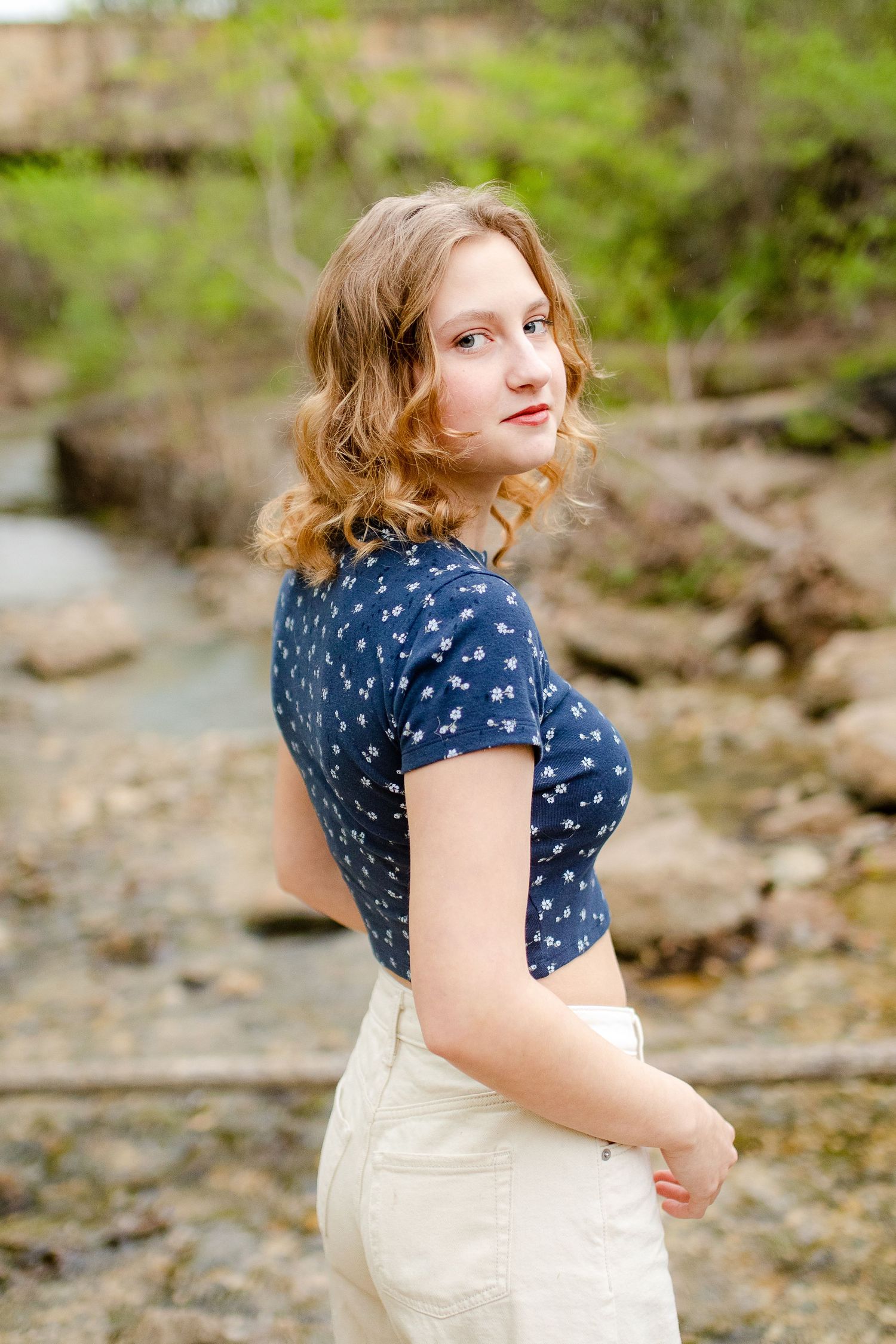Person in polka dot navy blouse and cream skirt poses by rocky stream in outdoor portrait setting.