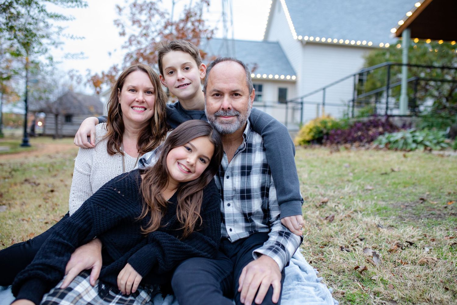 A family sitting together on a blanket outdoors with white house and autumn scenery in the background.