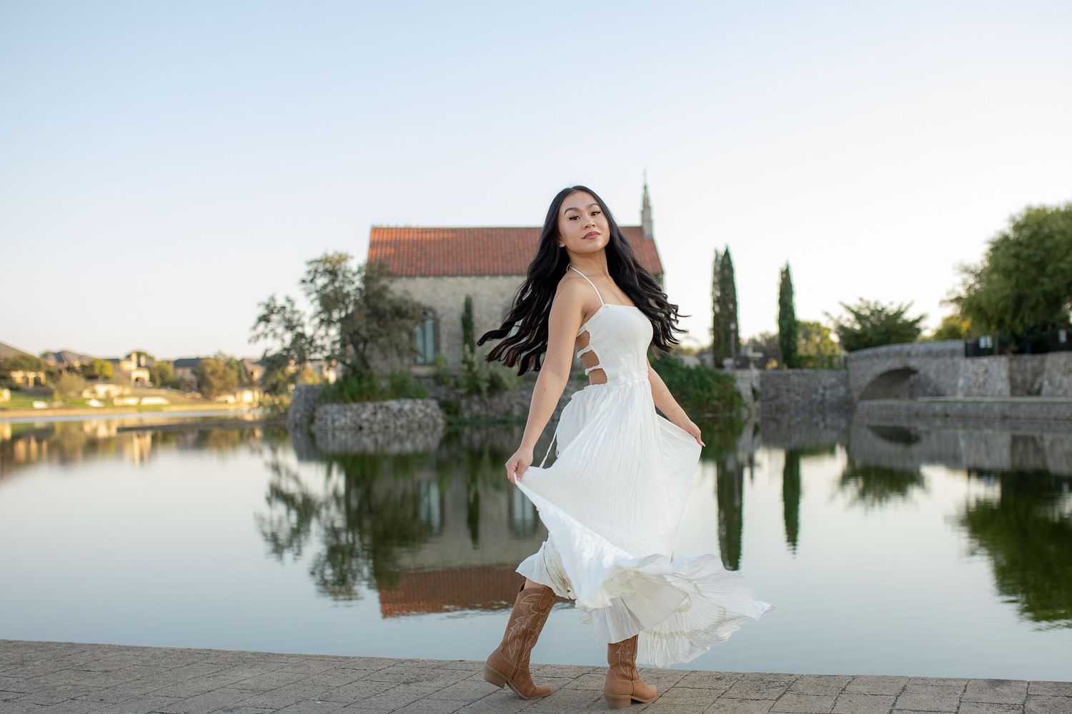 A joyful person twirls in a white dress by a serene lake with a rustic building and cypress trees reflected in the water.
