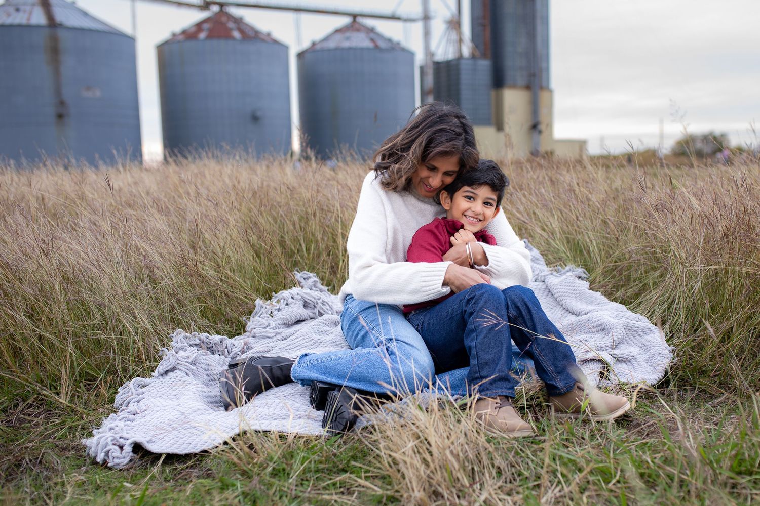 A tender moment captured on a blanket in tall grass with grain silos in the background.