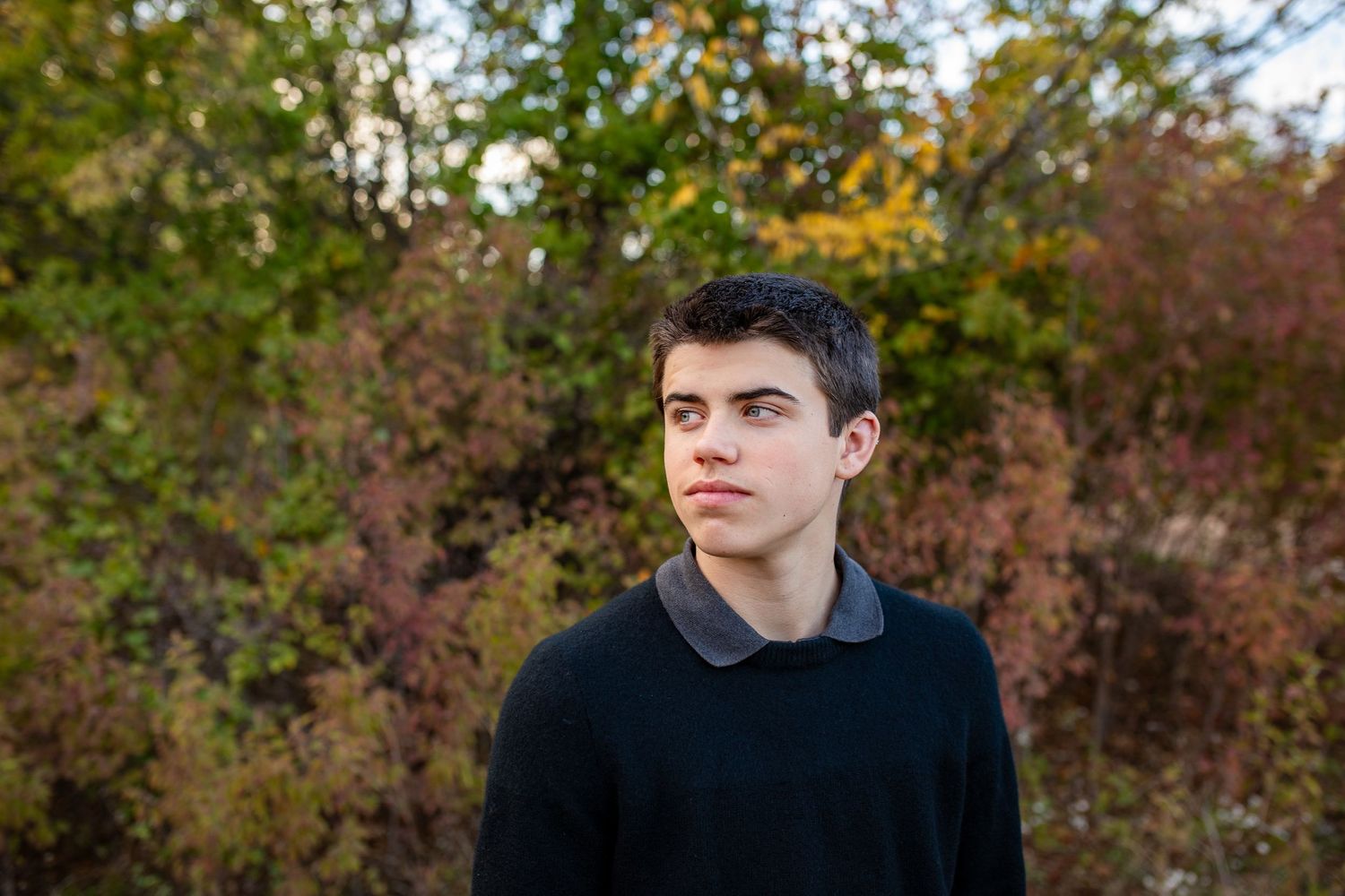 A young person in a black sweater stands outdoors against autumn foliage in natural lighting.