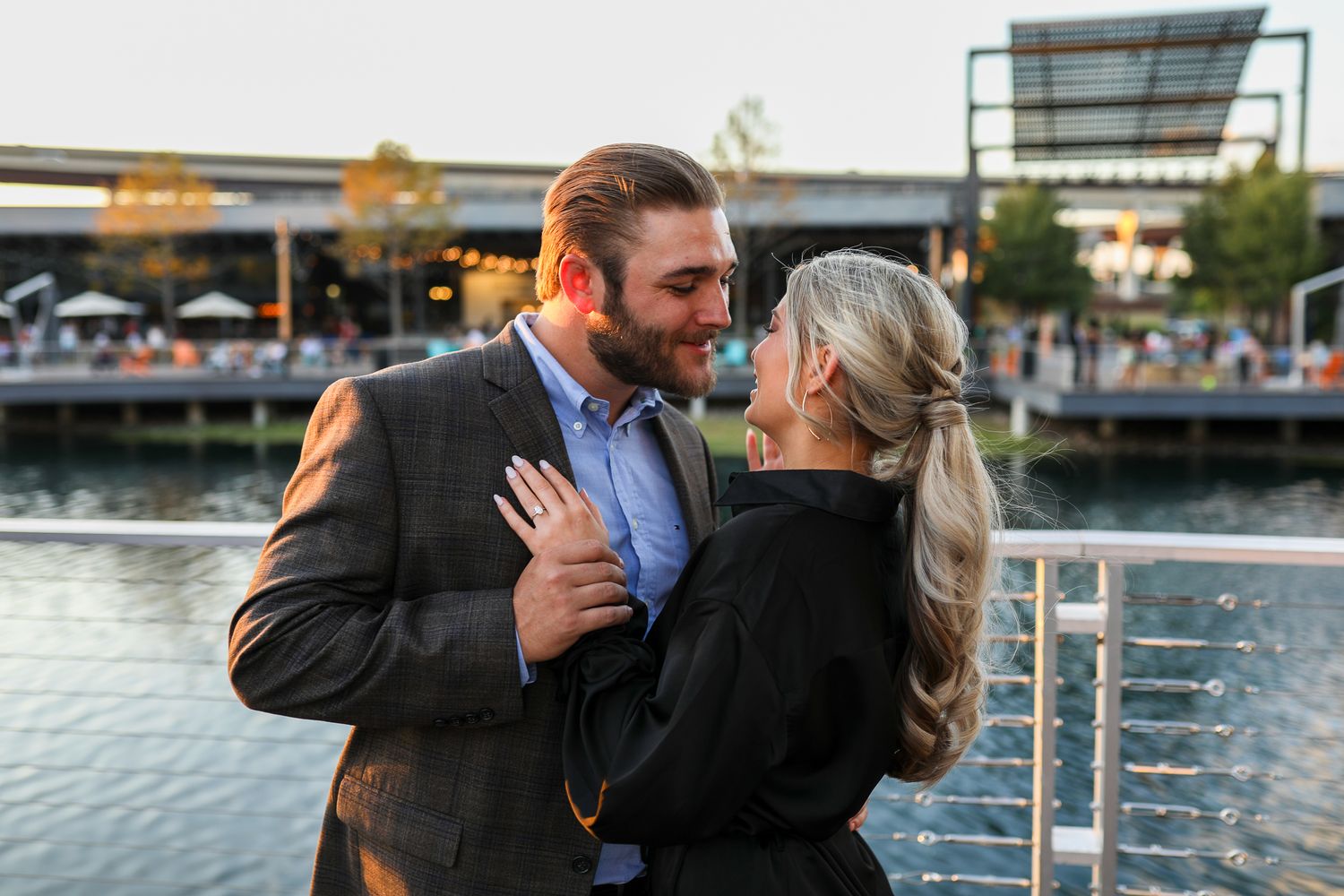 A couple in formal attire share an intimate moment on a waterfront deck during golden hour with a river view.