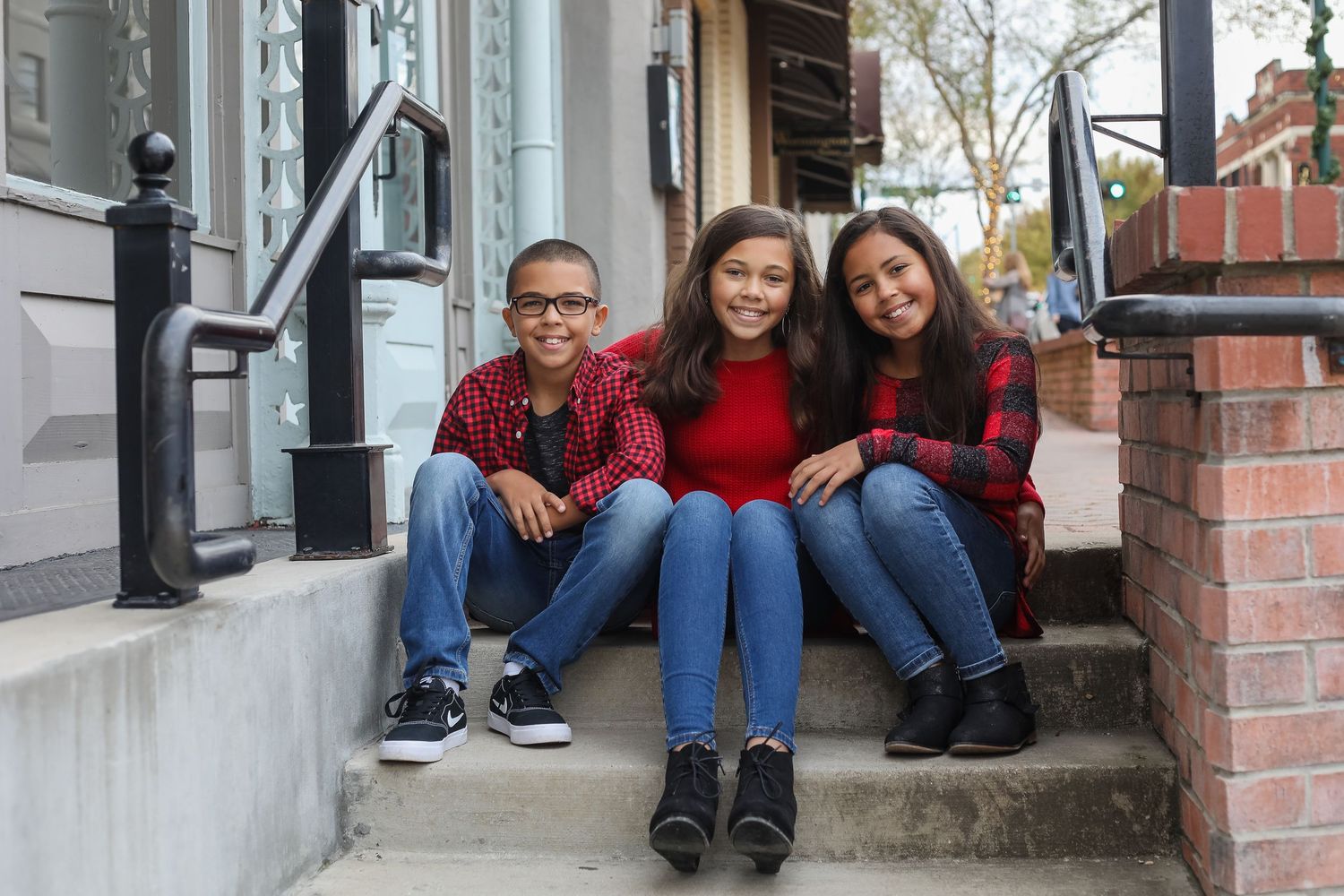 Three children in red and black plaid shirts sit together on outdoor steps smiling at the camera.