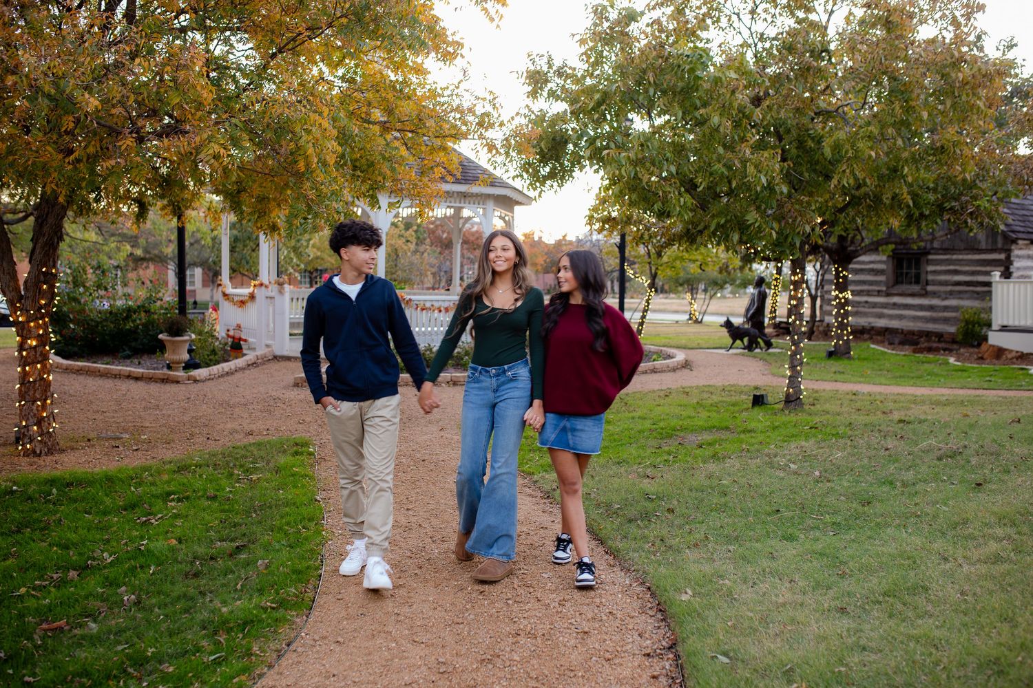 Three people walk together on a winding path through a garden with autumn trees and sunlight.