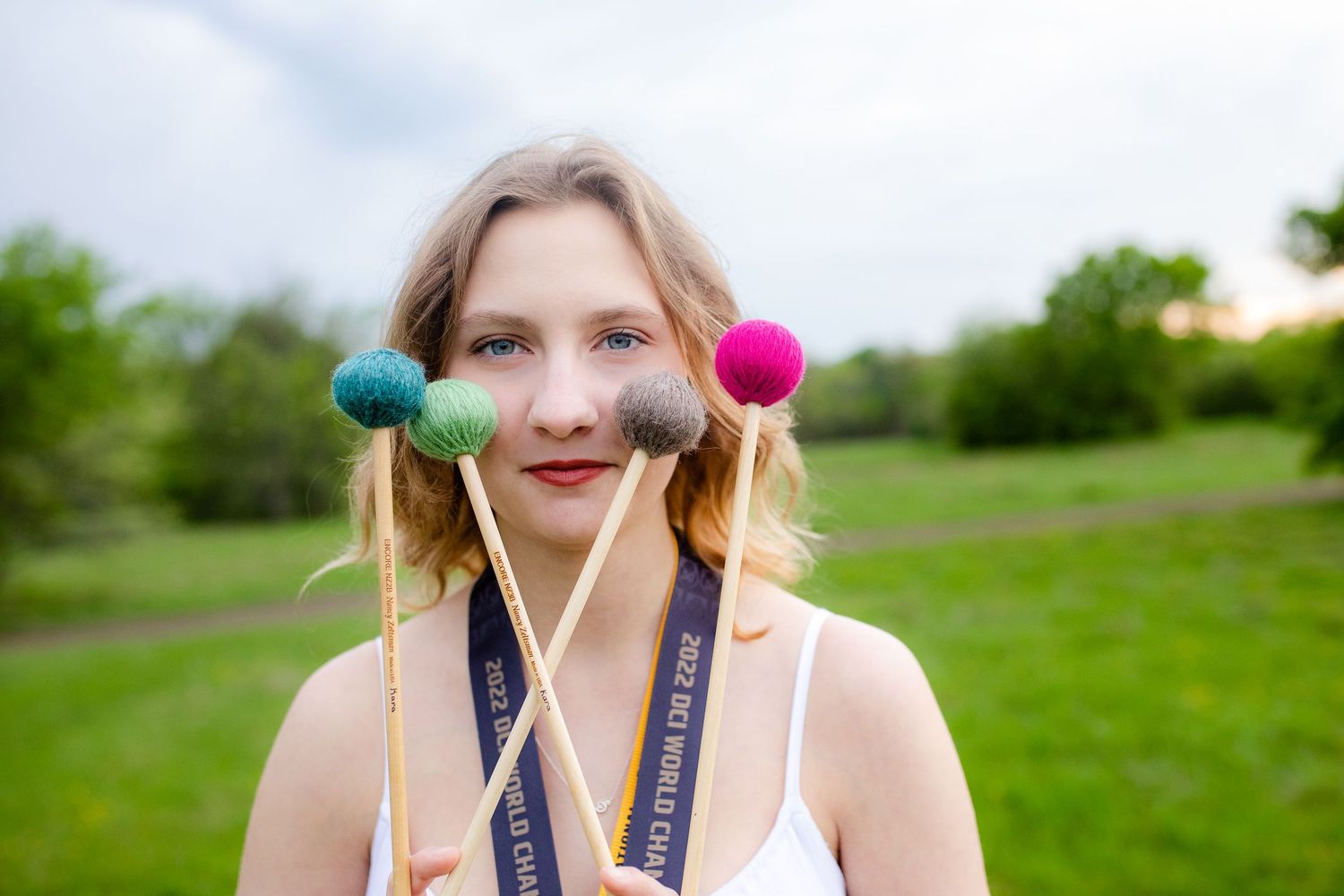 Person posing outdoors with colorful mallets and lanyard in a grassy park setting.