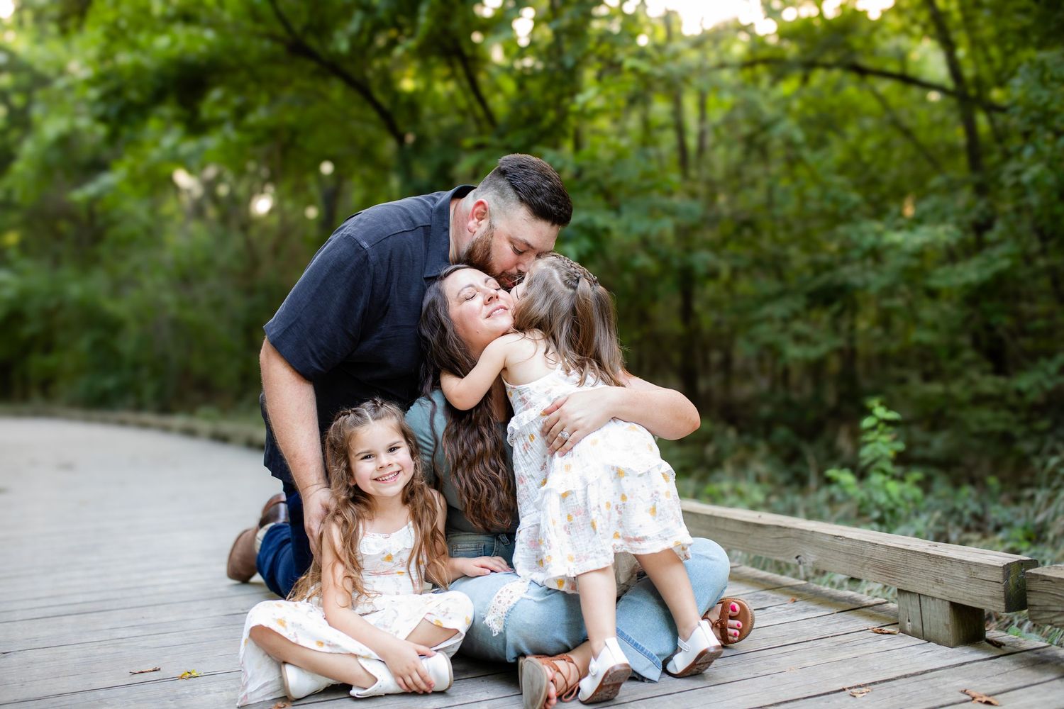 A family shares a tender moment together while sitting on a wooden bridge surrounded by lush greenery.