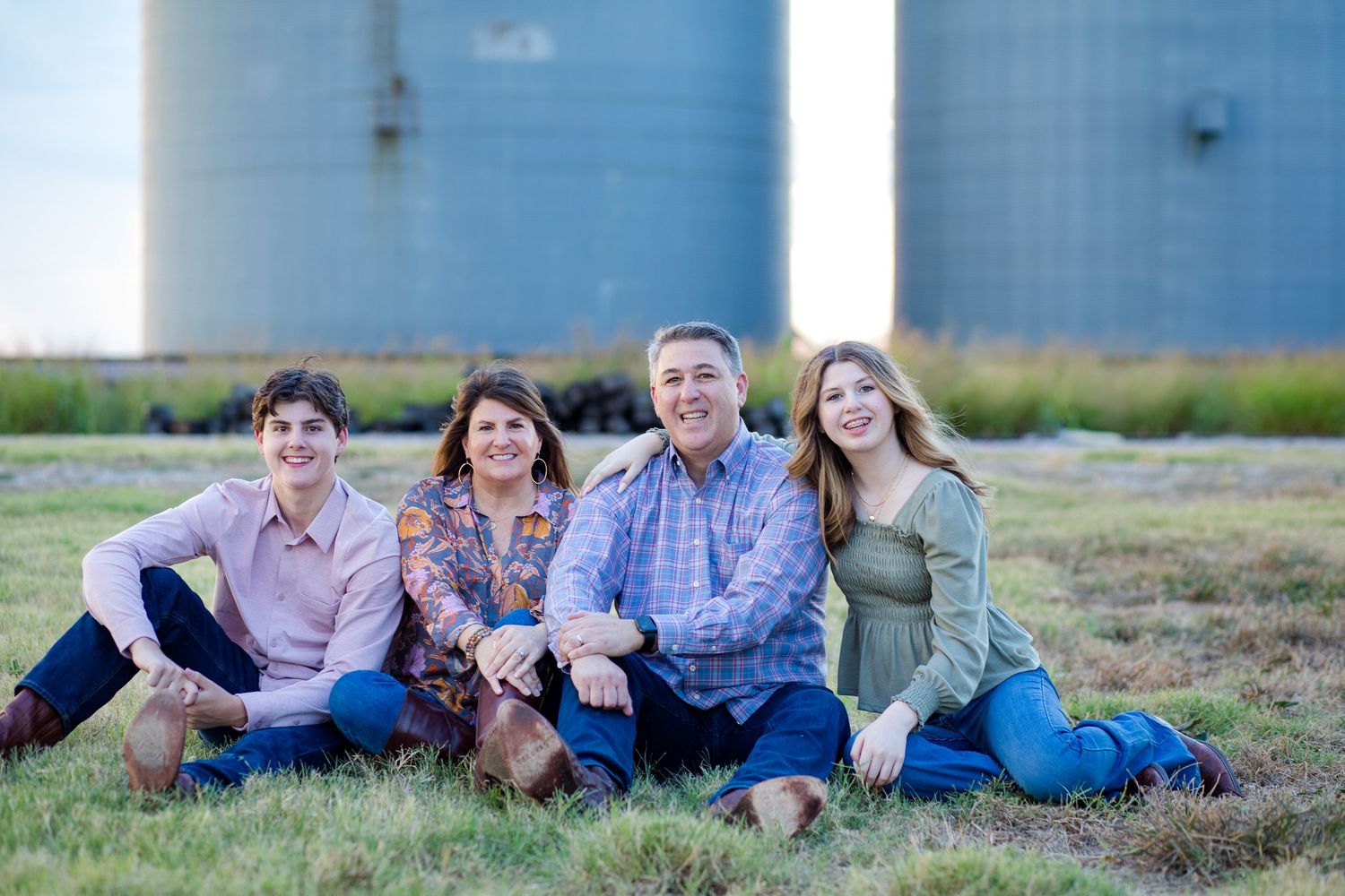 Four people sit together on grass in casual clothing with blue industrial tanks in the background.