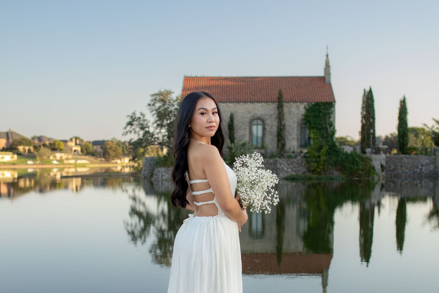 A person in a white dress holding baby's breath flowers stands beside a tranquil lake with a rustic church reflected in the water.