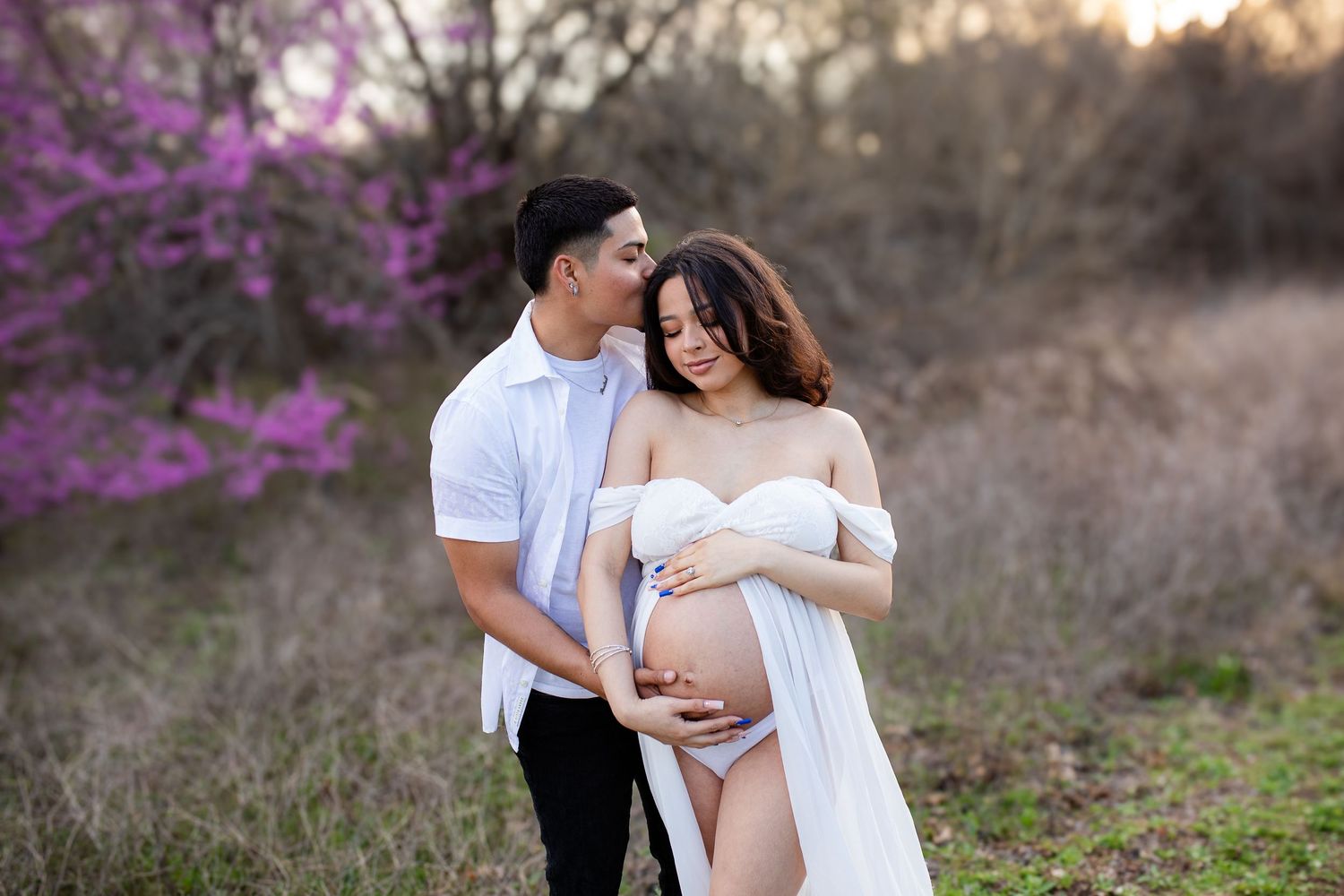 A couple in white embraces in a field with vibrant purple blossoming trees in the background at sunset.