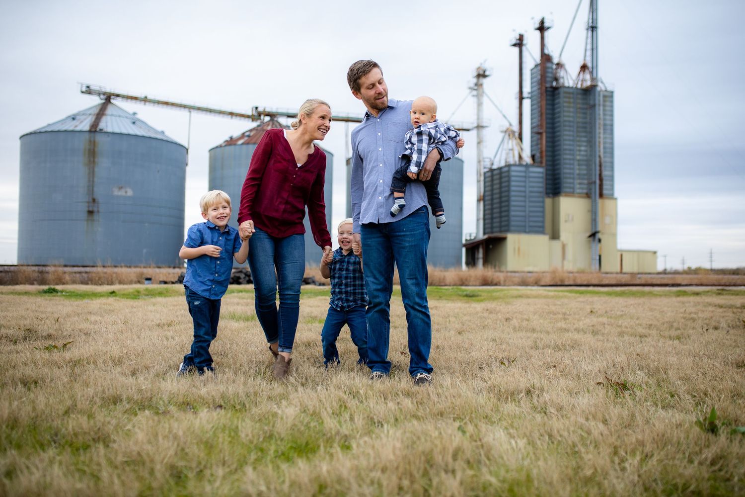 A family walks together in a grassy field with large industrial grain silos in the background.