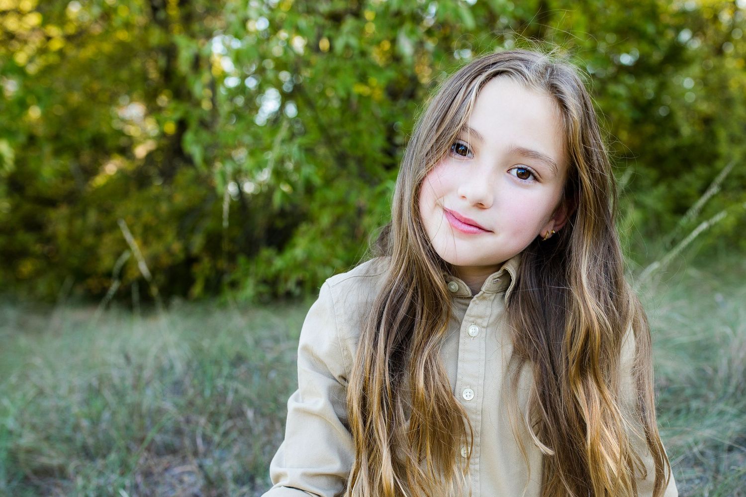 A person with long brown hair smiles while wearing a beige jacket outdoors with green foliage in the background.