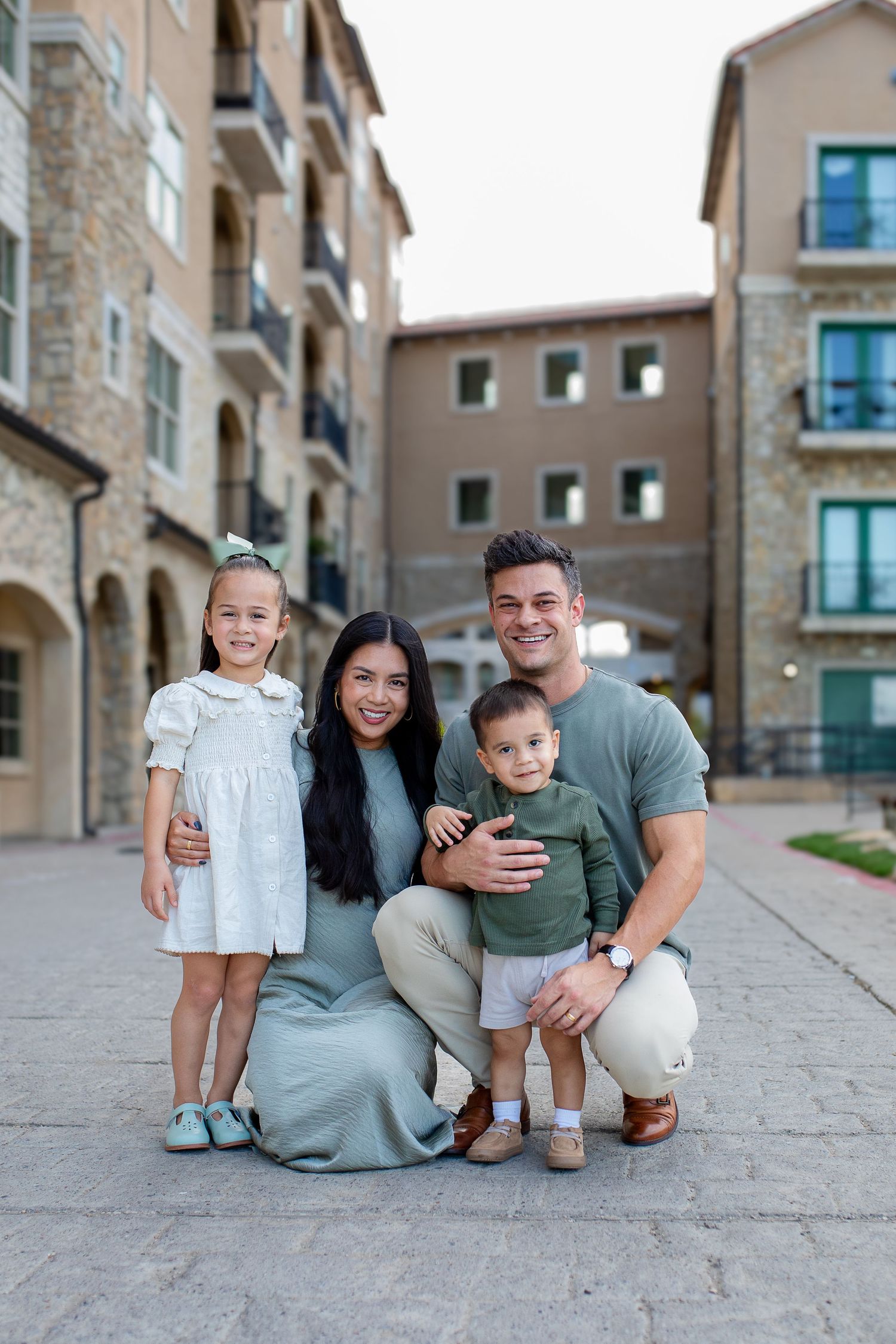 Family sitting and playing together on stone pavement outside modern residential buildings on sunny day.