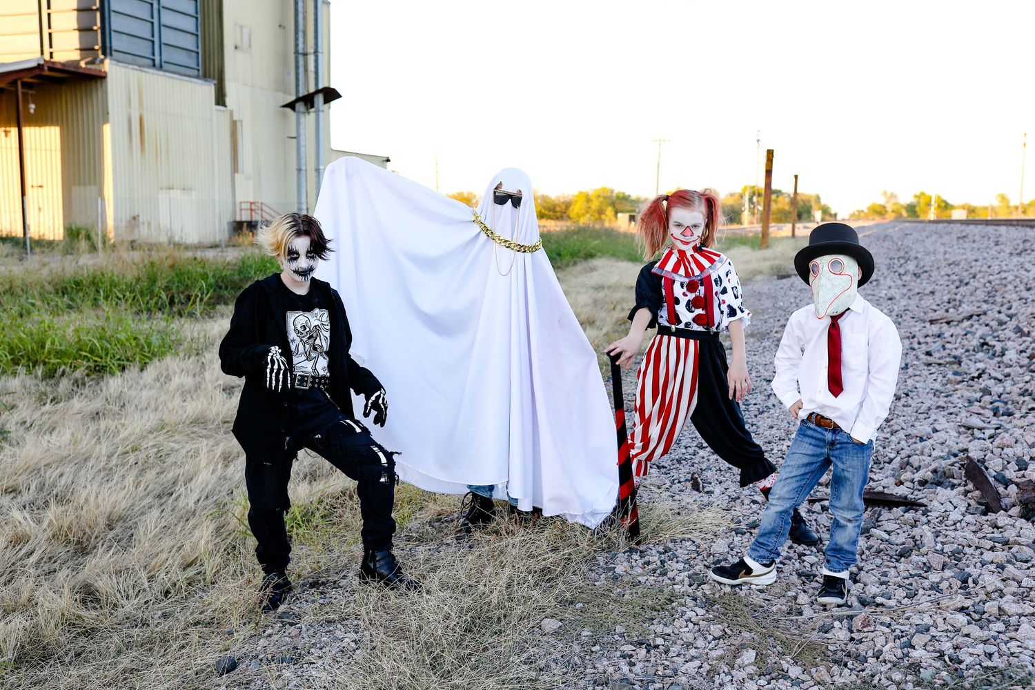 Four festive figures in Halloween costumes pose outdoors near a building at sunset.