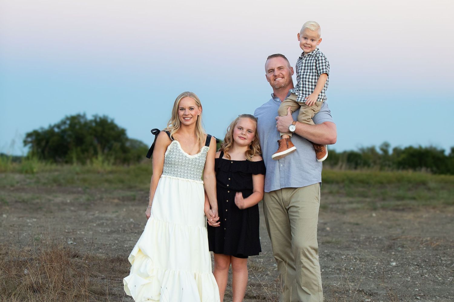 A family in matching casual outfits stands together smiling on an open field at sunset.