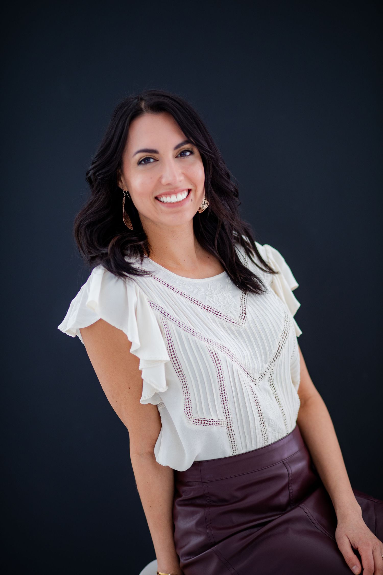 Professional portrait against dark background showing a white ruffled blouse paired with a burgundy pencil skirt.