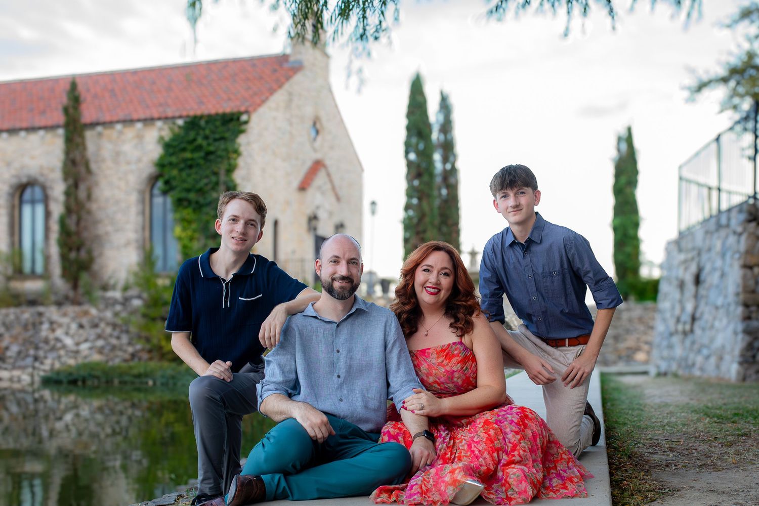 A group of four people sitting casually together in front of a rustic stone building with cypress trees and ivy.