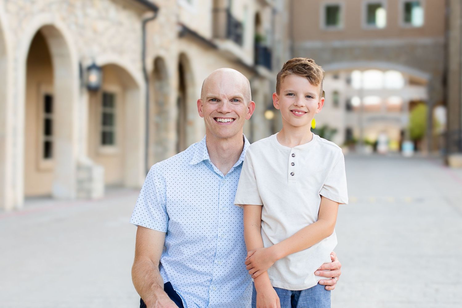 Two people stand together smiling outdoors in front of a beige building with arched doorways on a sunny day.