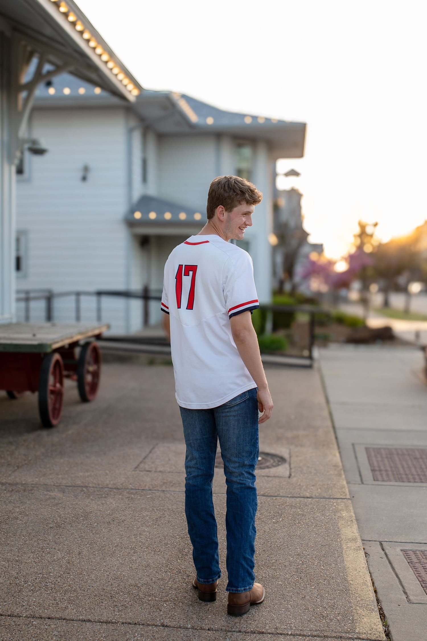 Person wearing white jersey number 17 and jeans stands on a sidewalk at sunset near a historic building.