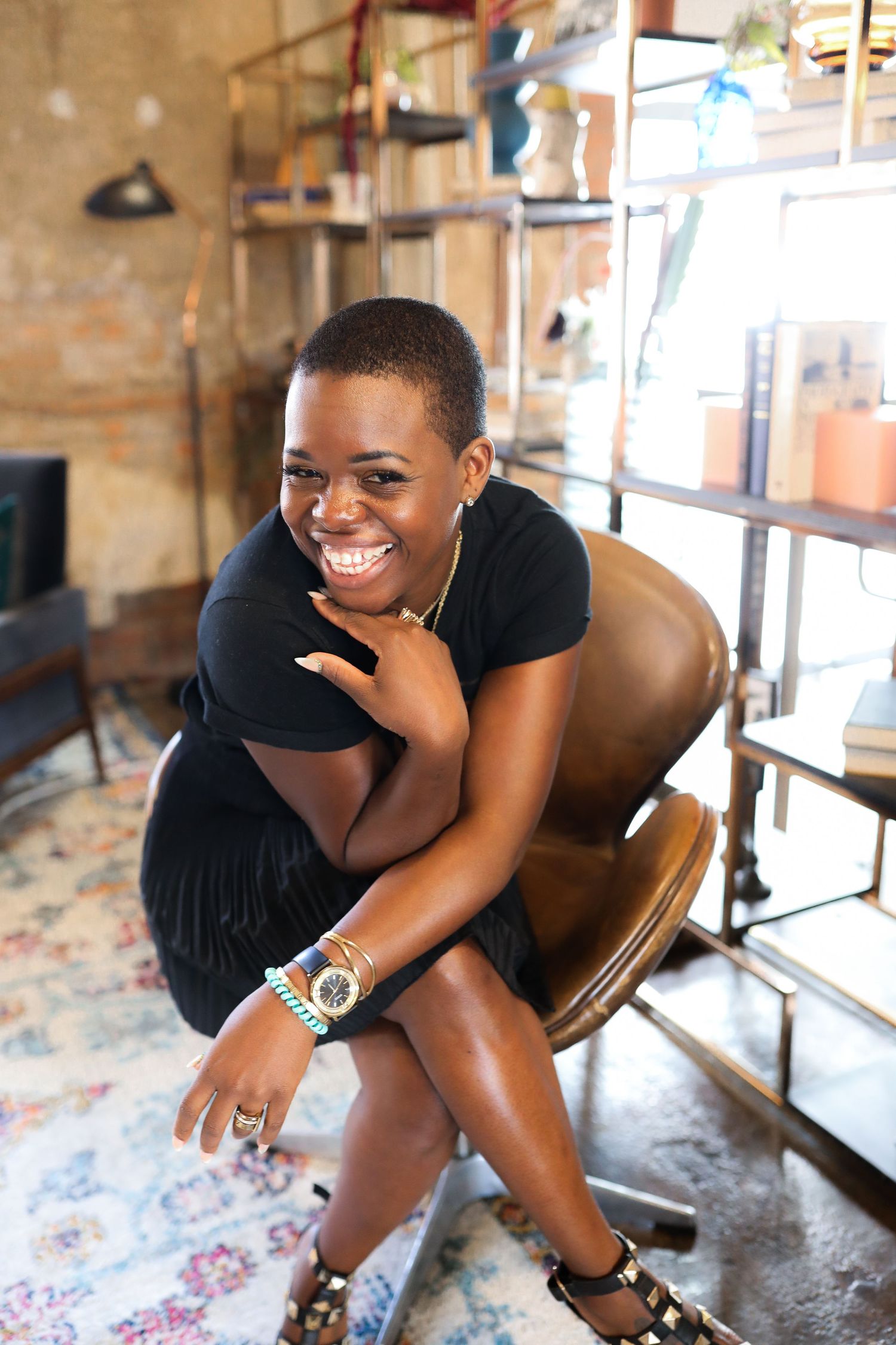 Someone wearing a black outfit sits casually in a brown chair with a bright smile while leaning forward.