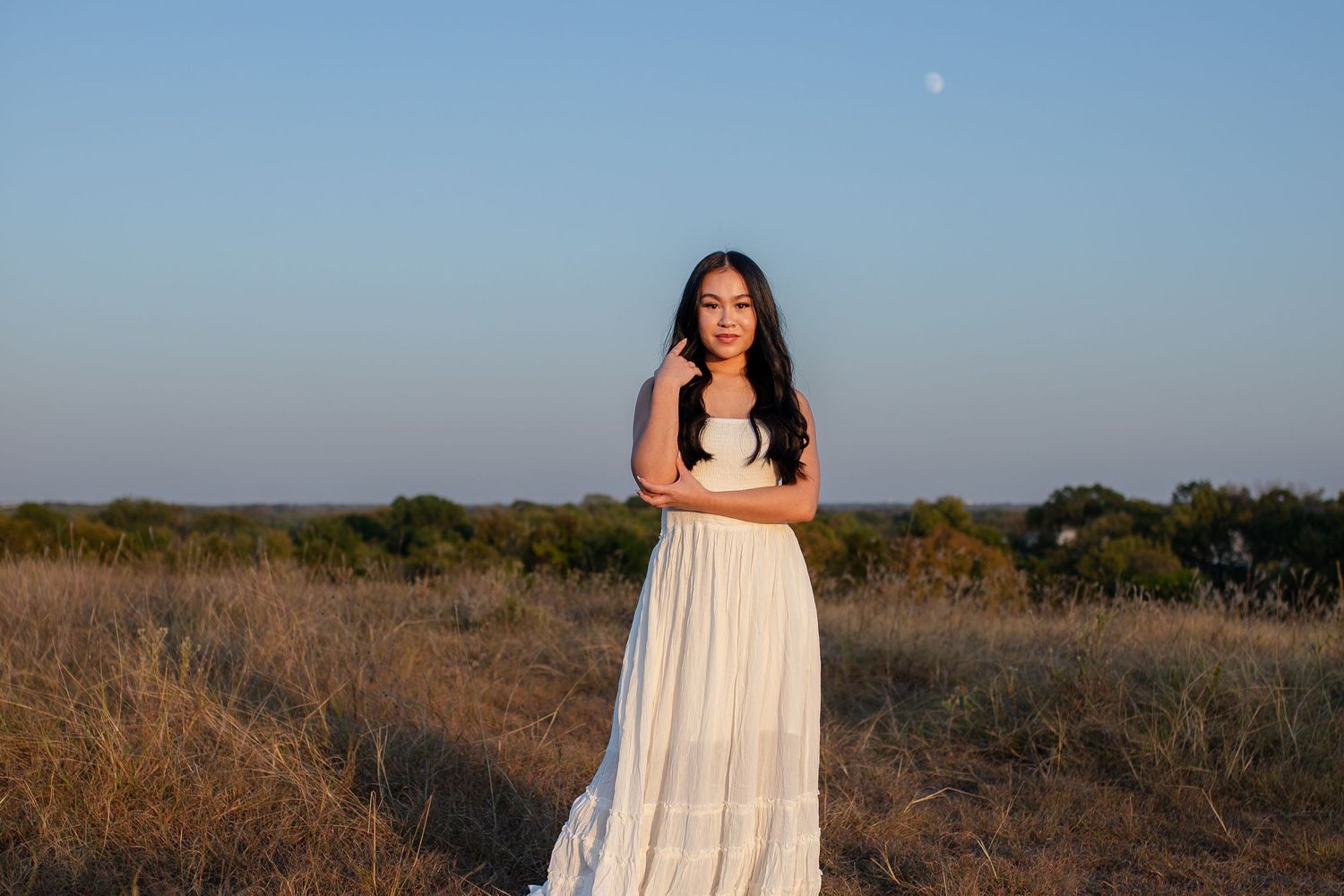 A person stands in a white flowing dress against a scenic rural landscape at golden hour.