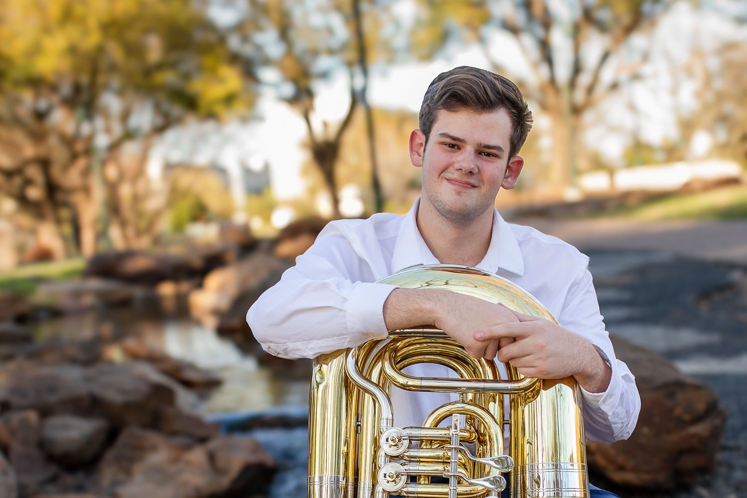 Musician in white shirt holds brass tuba instrument outdoors with autumn foliage in background.