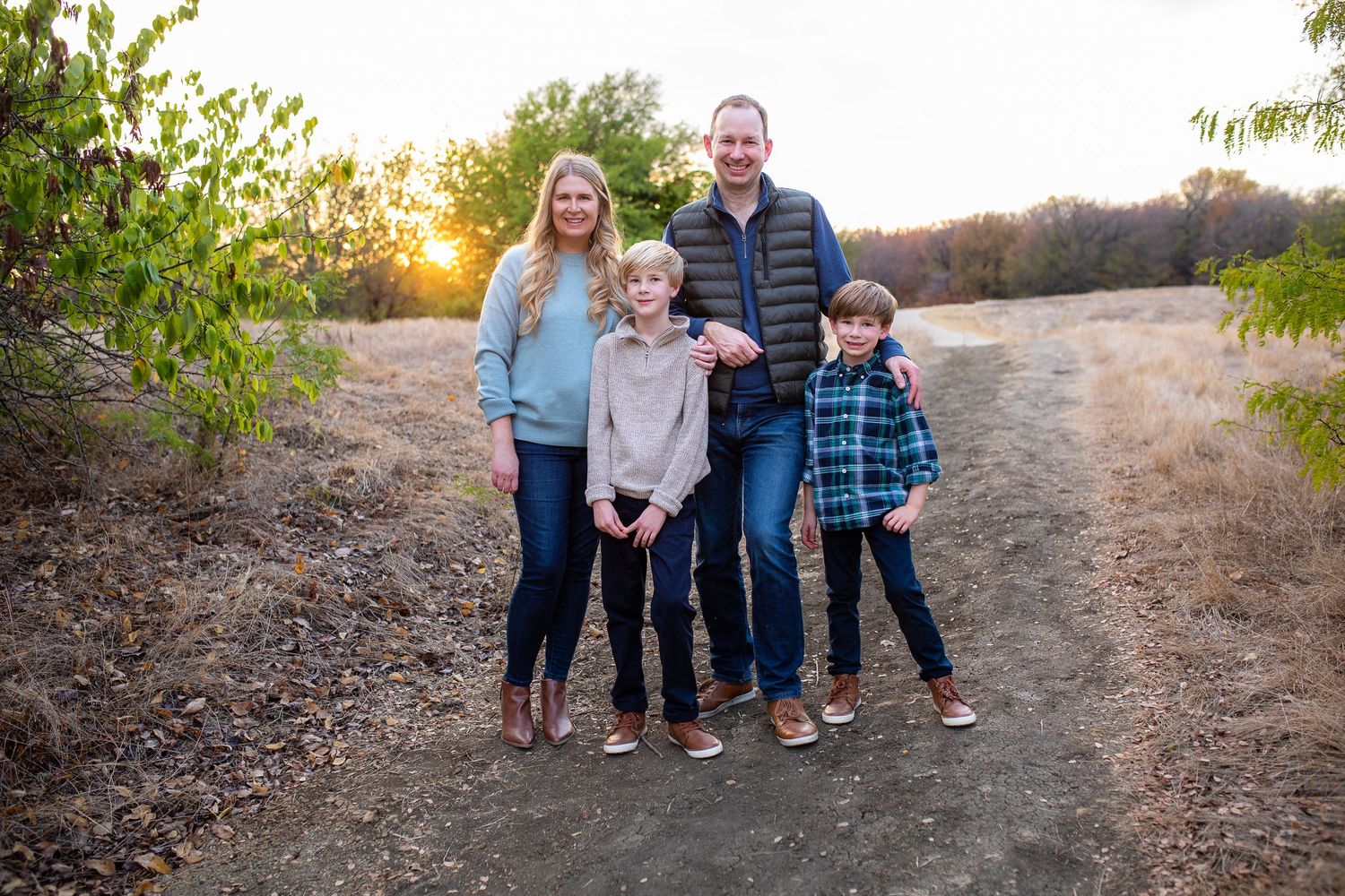 A family of four stands together on a dirt path at sunset, surrounded by green foliage.