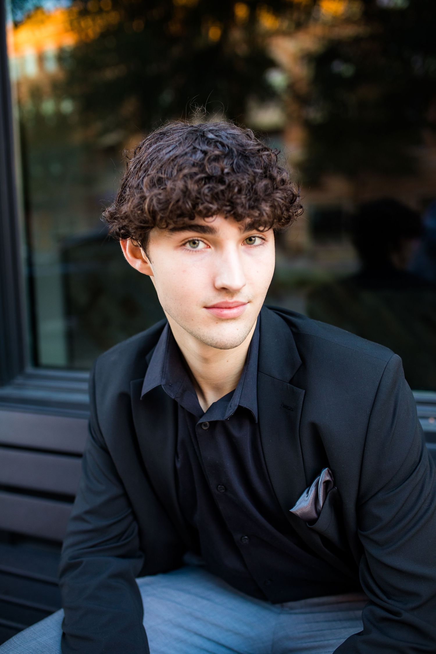 Portrait of a young person in all black formal attire with curly dark hair sitting outdoors at dusk.