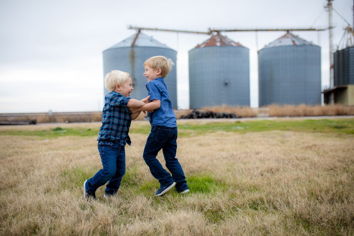 Two young children in blue outfits play together in a grassy field with grain silos in the background.