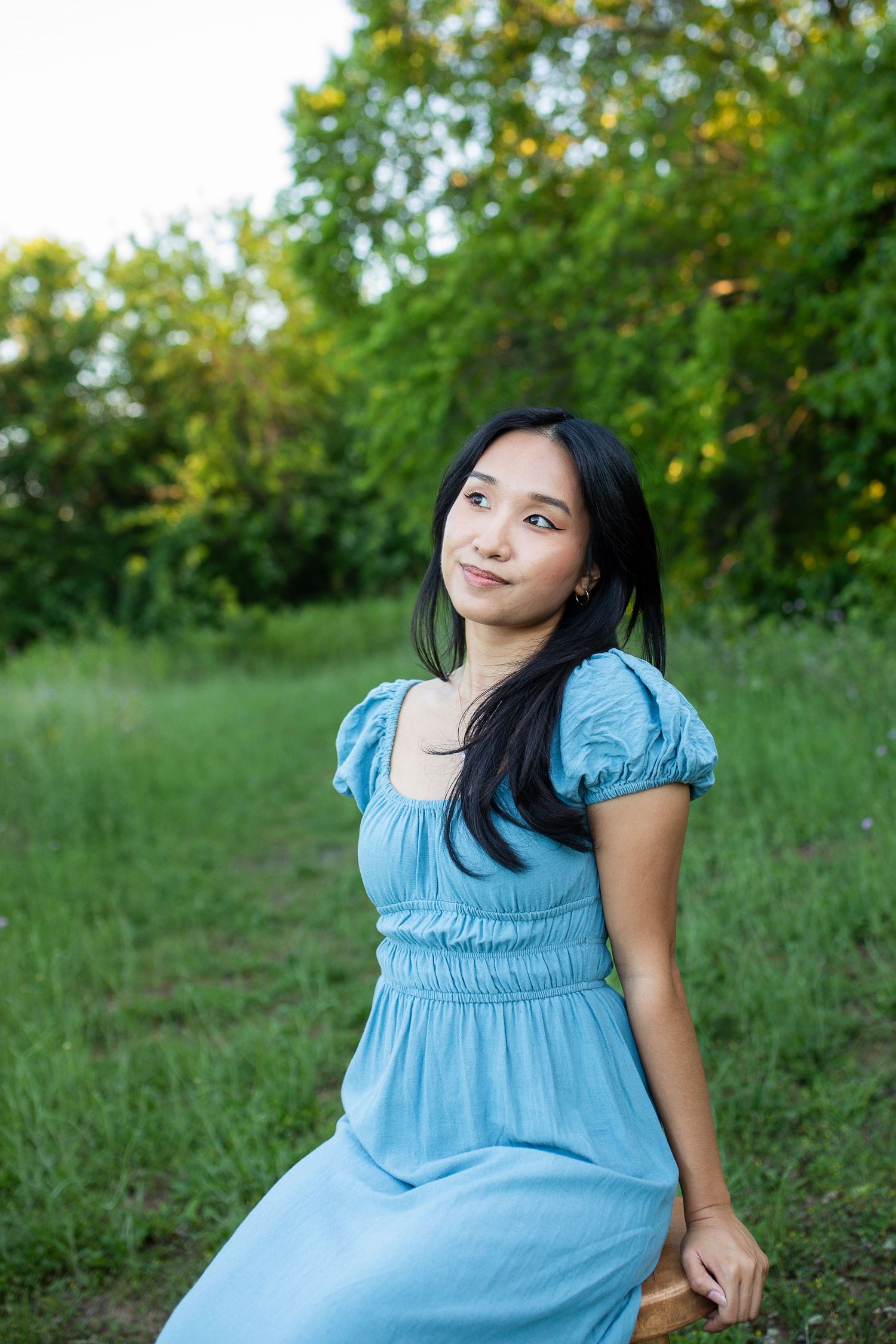 A woman in a light blue summer dress sits outdoors in a sunlit grassy area surrounded by green foliage.