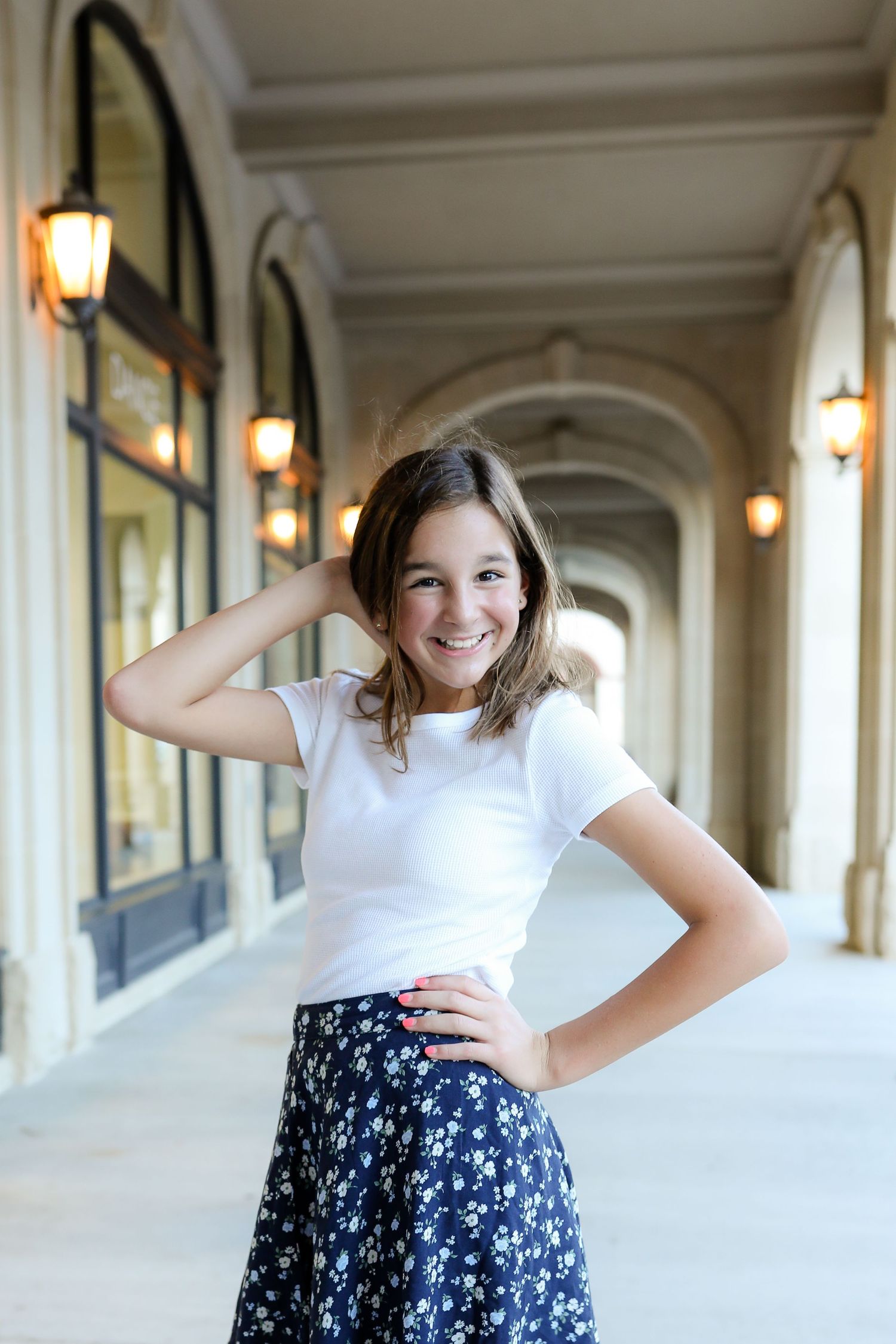 A young person in a white shirt and navy polka dot skirt poses playfully in an elegant hallway with arched columns.