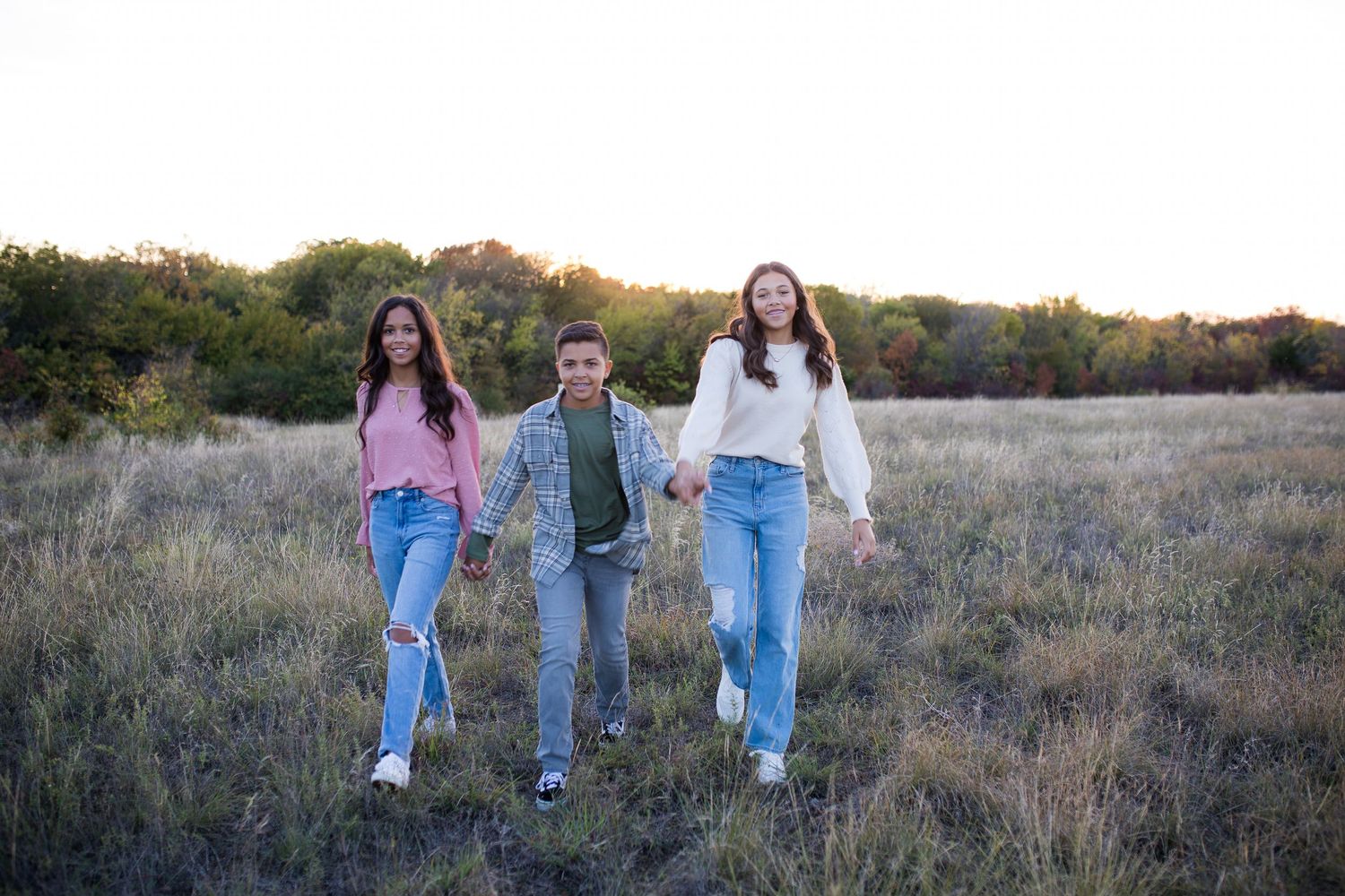 Three young friends holding hands while walking together through a scenic autumn field at sunset.
