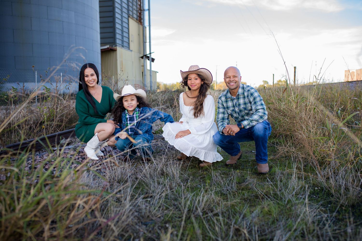 A seated family in casual clothing poses together on a grassy hill during sunset with the sky in the background.