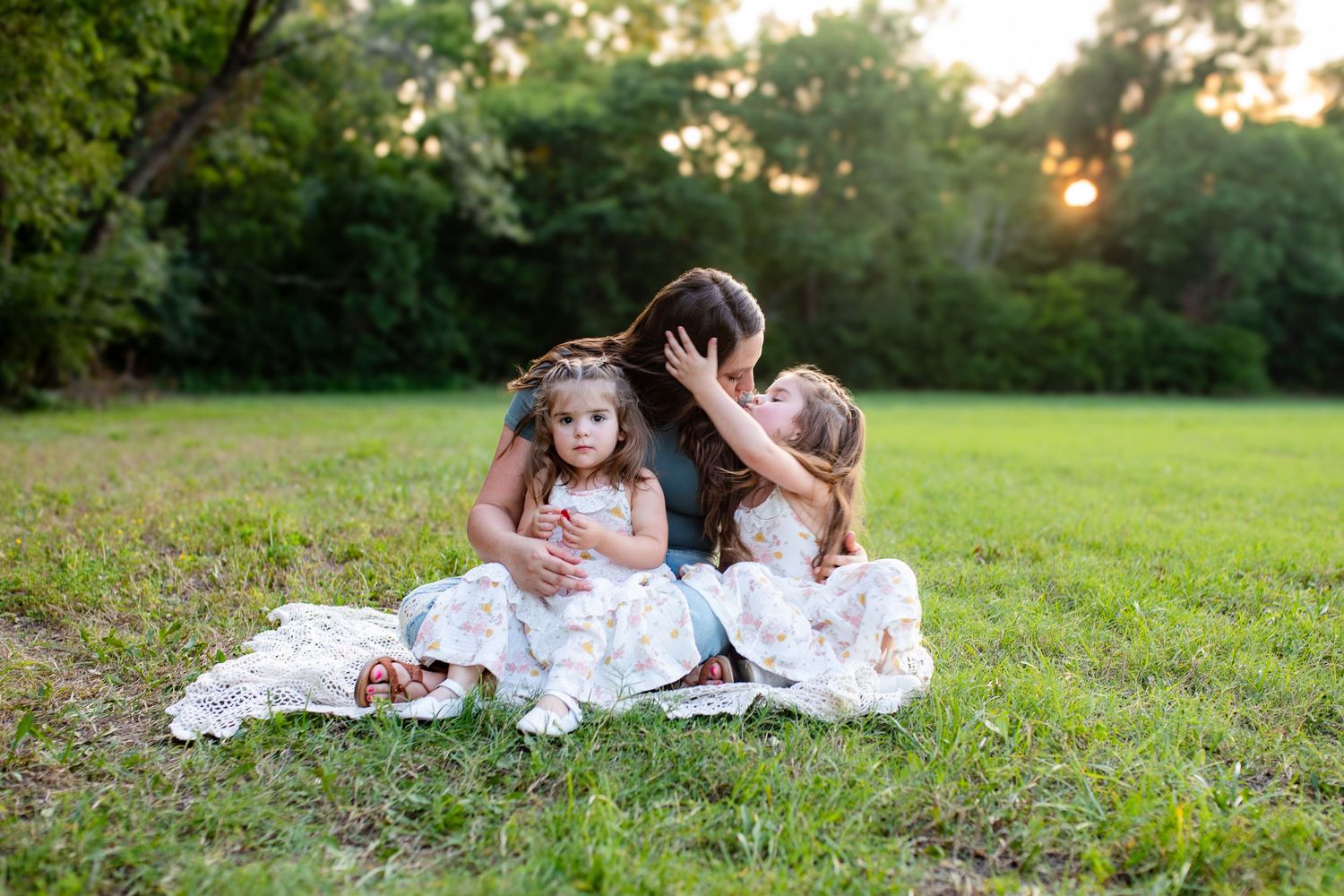 Three people in white lace dresses sit together on a blanket in a grassy field at sunset.