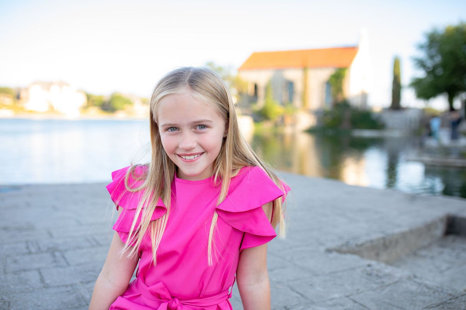 Young person in bright pink dress sitting by waterfront with buildings and boats in background.