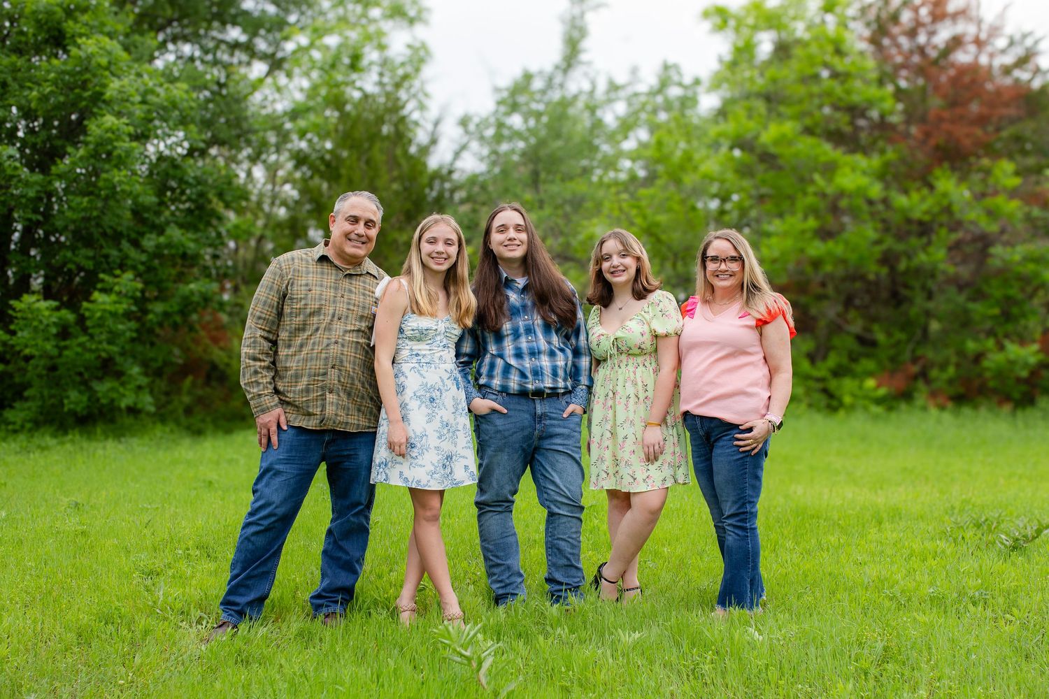 A group of people stands together smiling in a green grassy field with trees in the background on a sunny day.