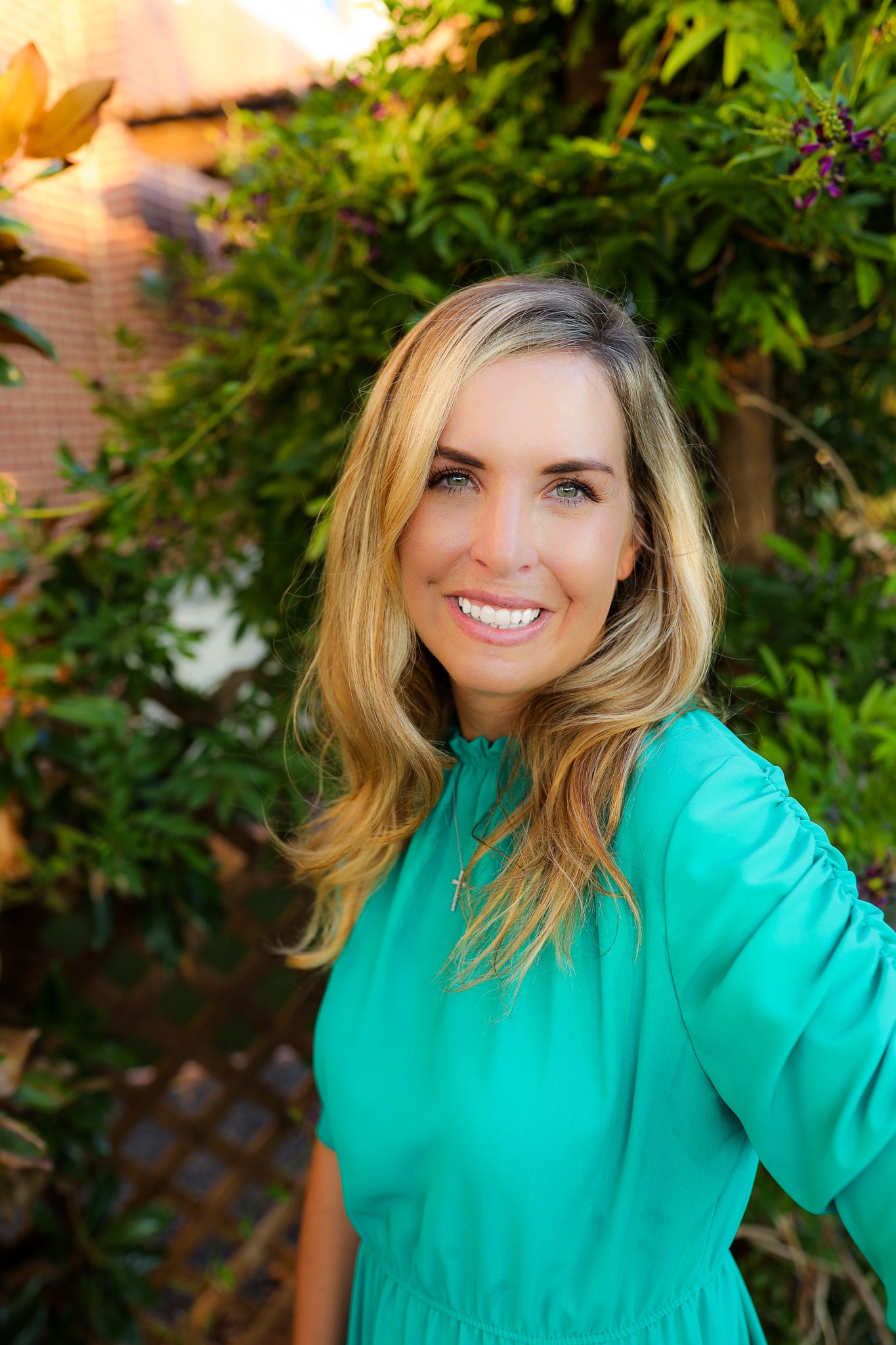 A woman in a vibrant turquoise dress poses outdoors in front of lush green foliage at golden hour.