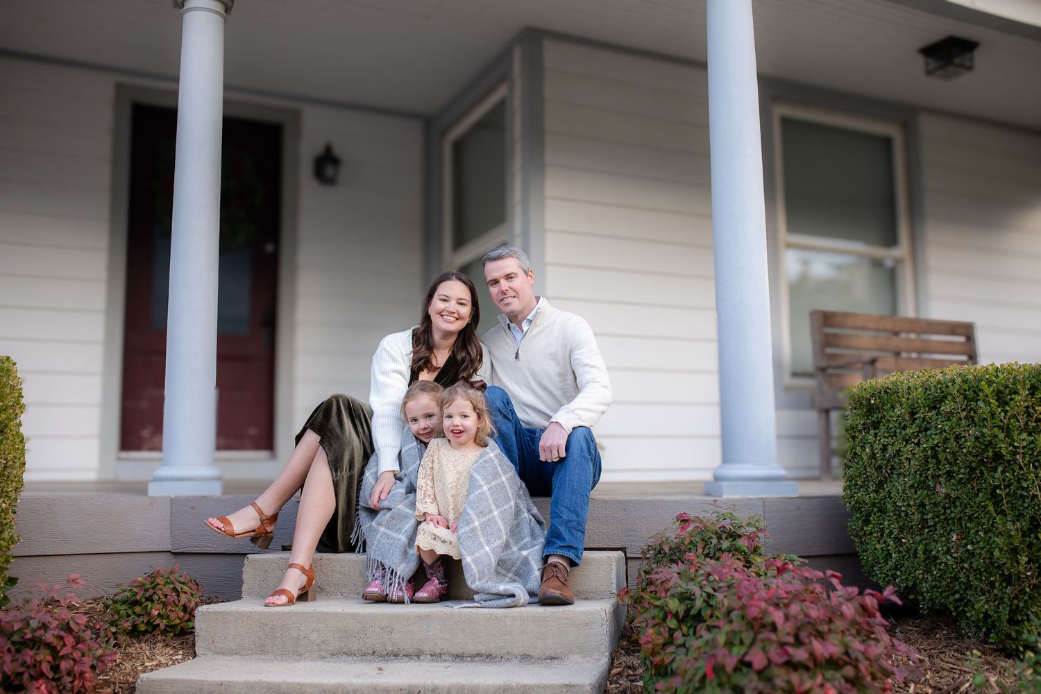 Three people sit relaxed together on front porch steps of a craftsman style house with white pillars and pink flowers.