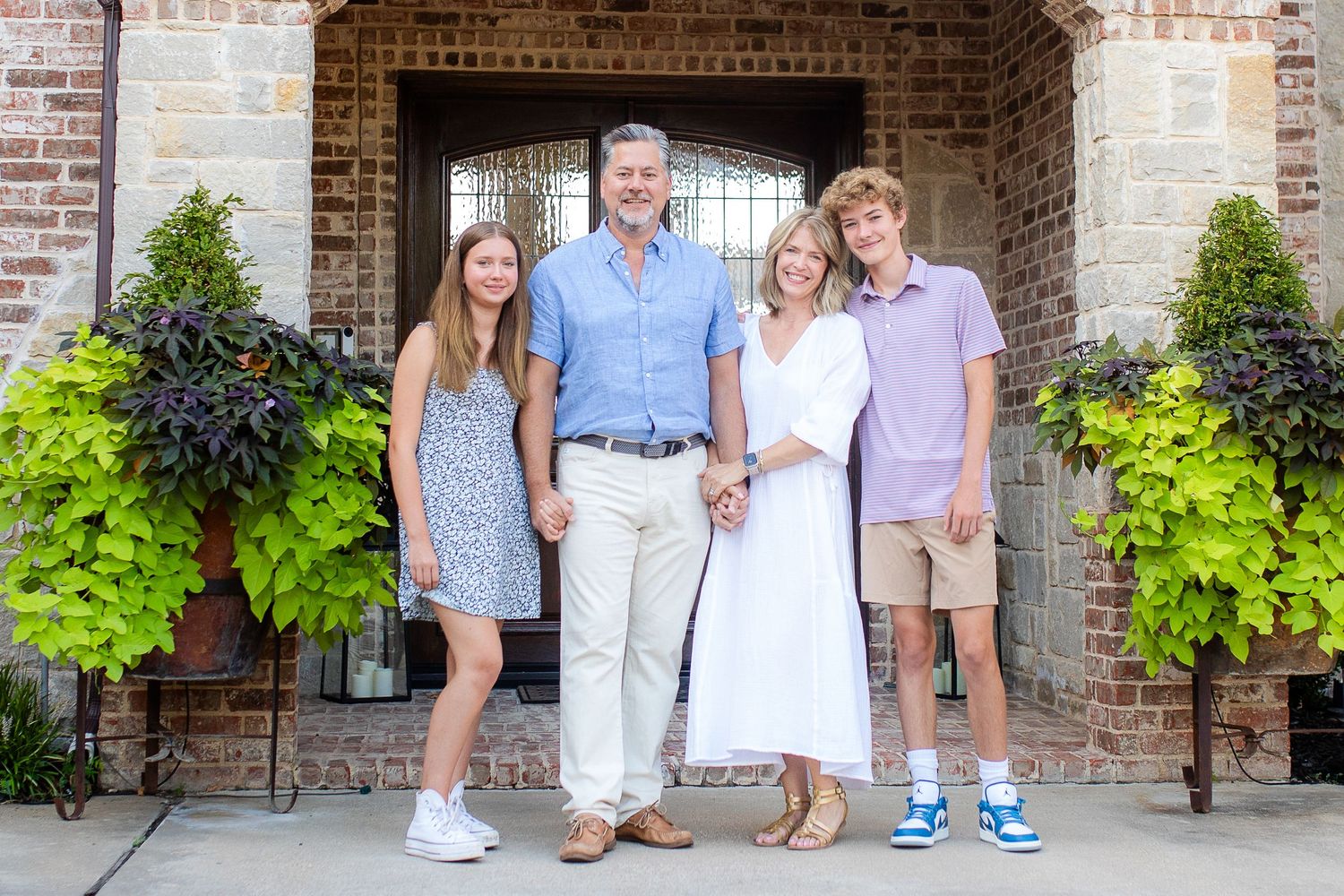 A family stands together on a brick porch with decorative potted plants on either side during a summer photo session.
